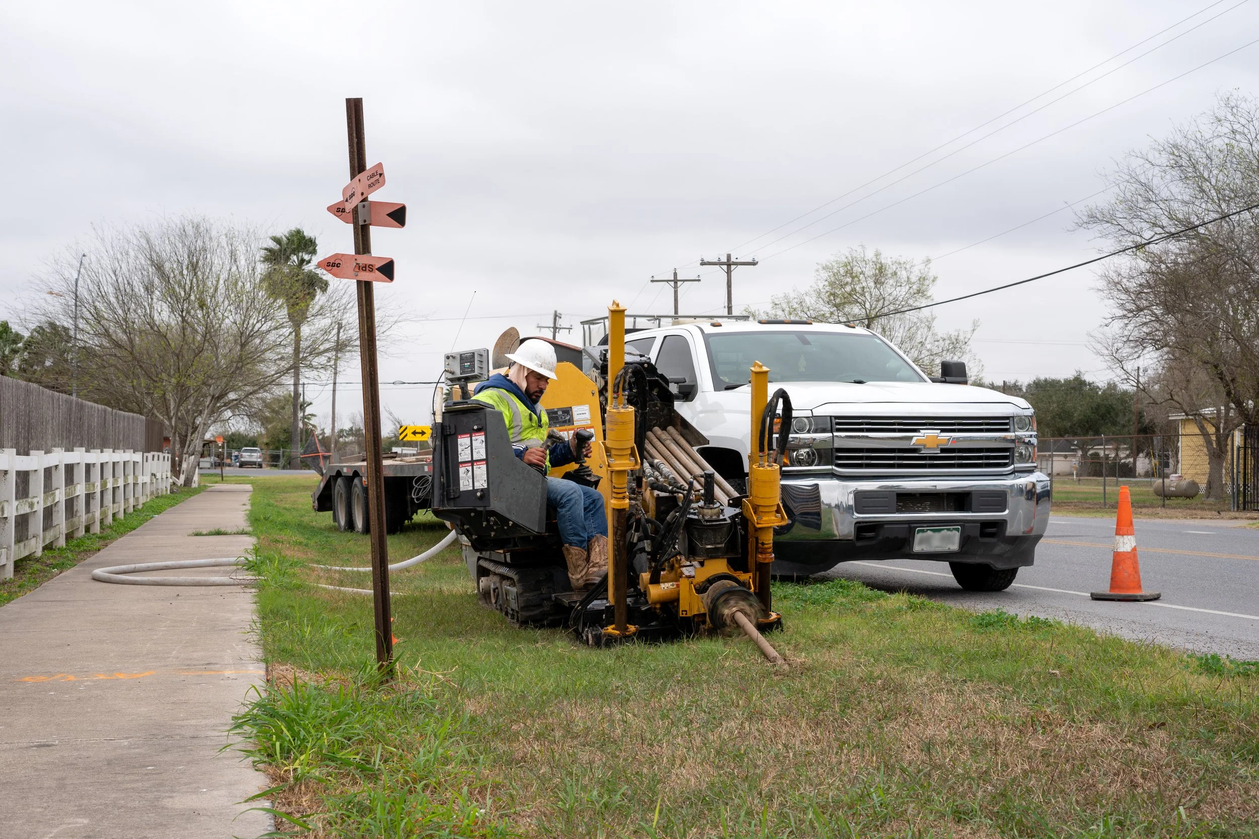 A construction worker in a white hard hat and reflective vest operating machinery beside a white Chevrolet truck on a grassy roadside during cloudy weather.