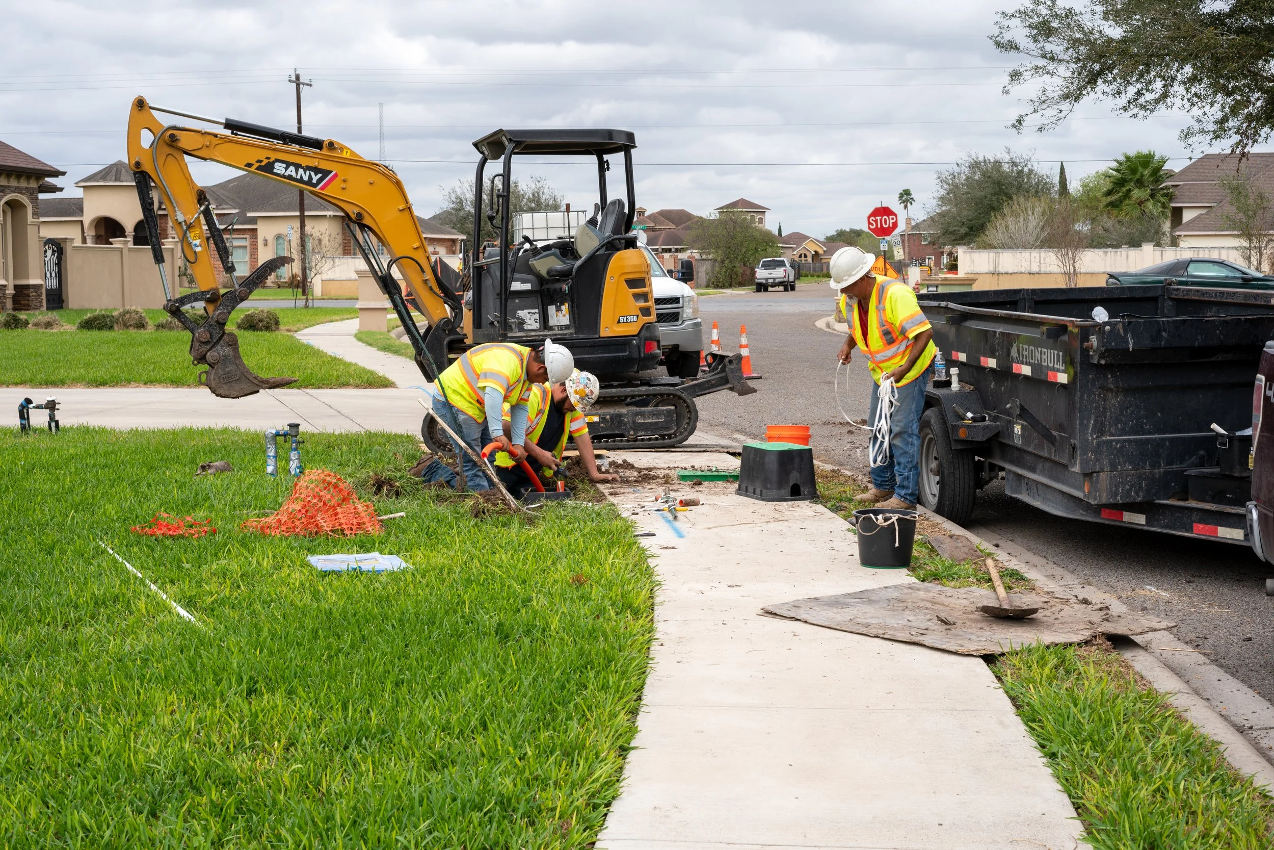 Construction workers installing or repairing sidewalk near curb on a residential street with houses, trees, and vehicles in the background.