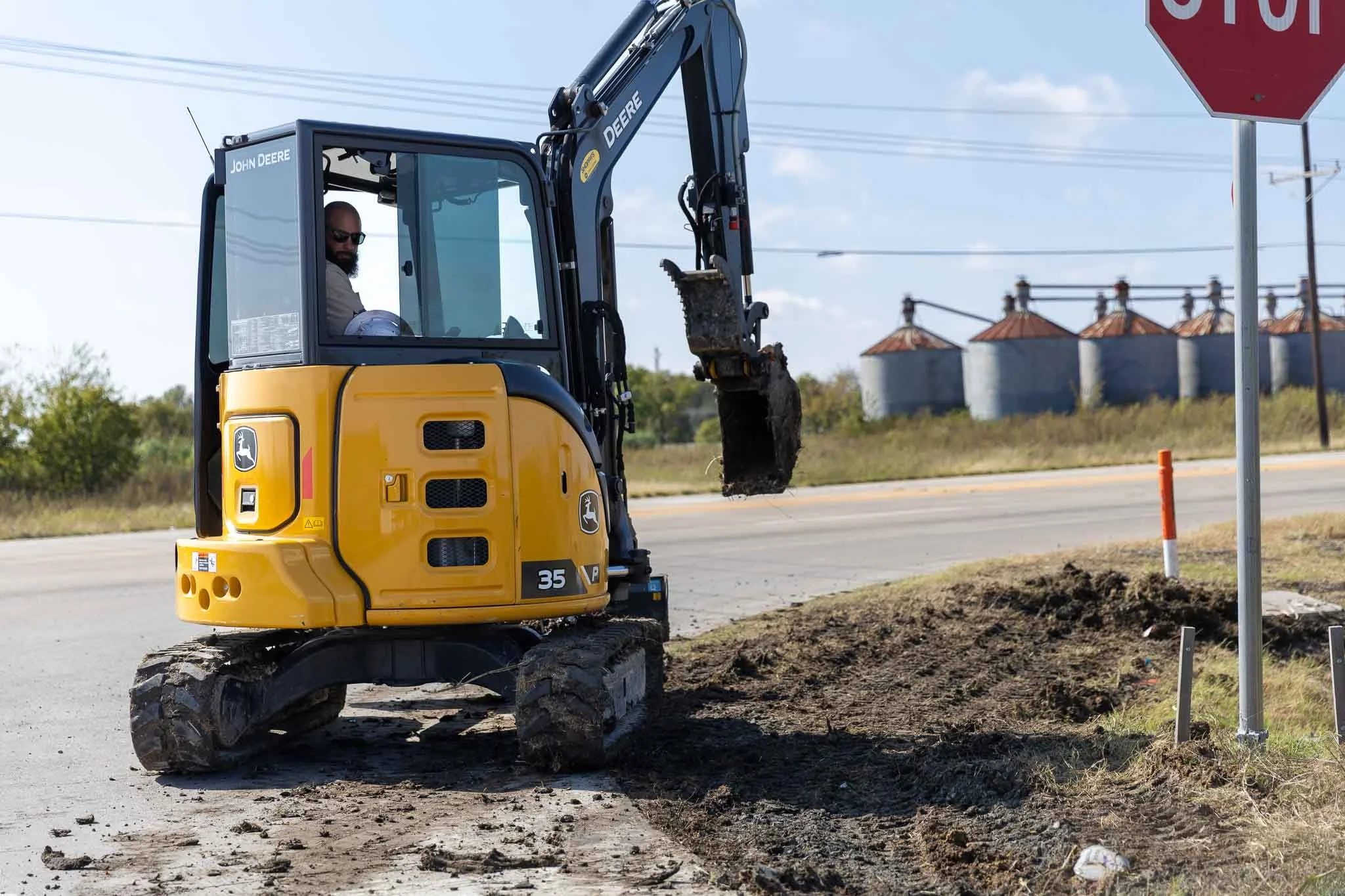 A man operating a yellow John Deere compact excavator near a roadside stop sign, digging into the earth during daytime under a partly cloudy sky.