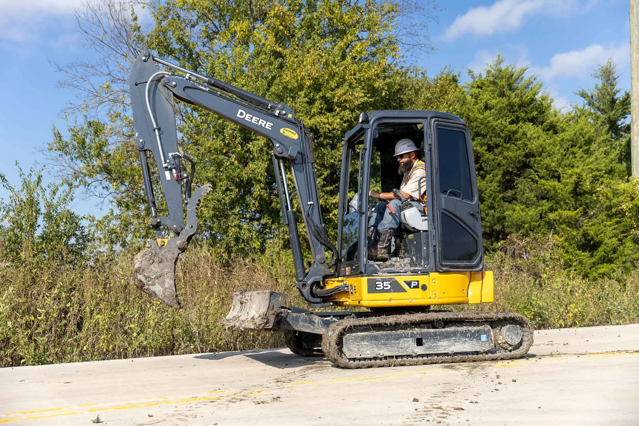 A construction worker operates a small John Deere excavator on a construction site, with trees and blue sky in the background.