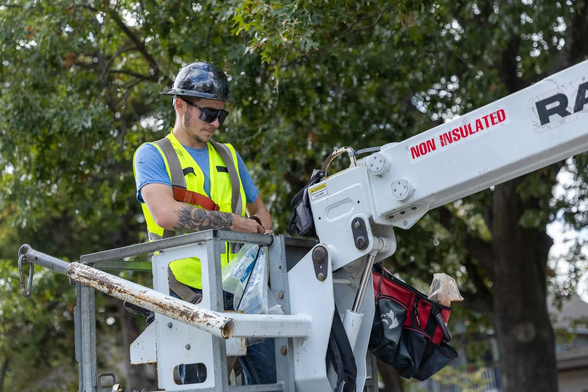 A male construction worker wearing a safety vest, helmet, and sunglasses is working on a lift while outdoors with trees in the background.