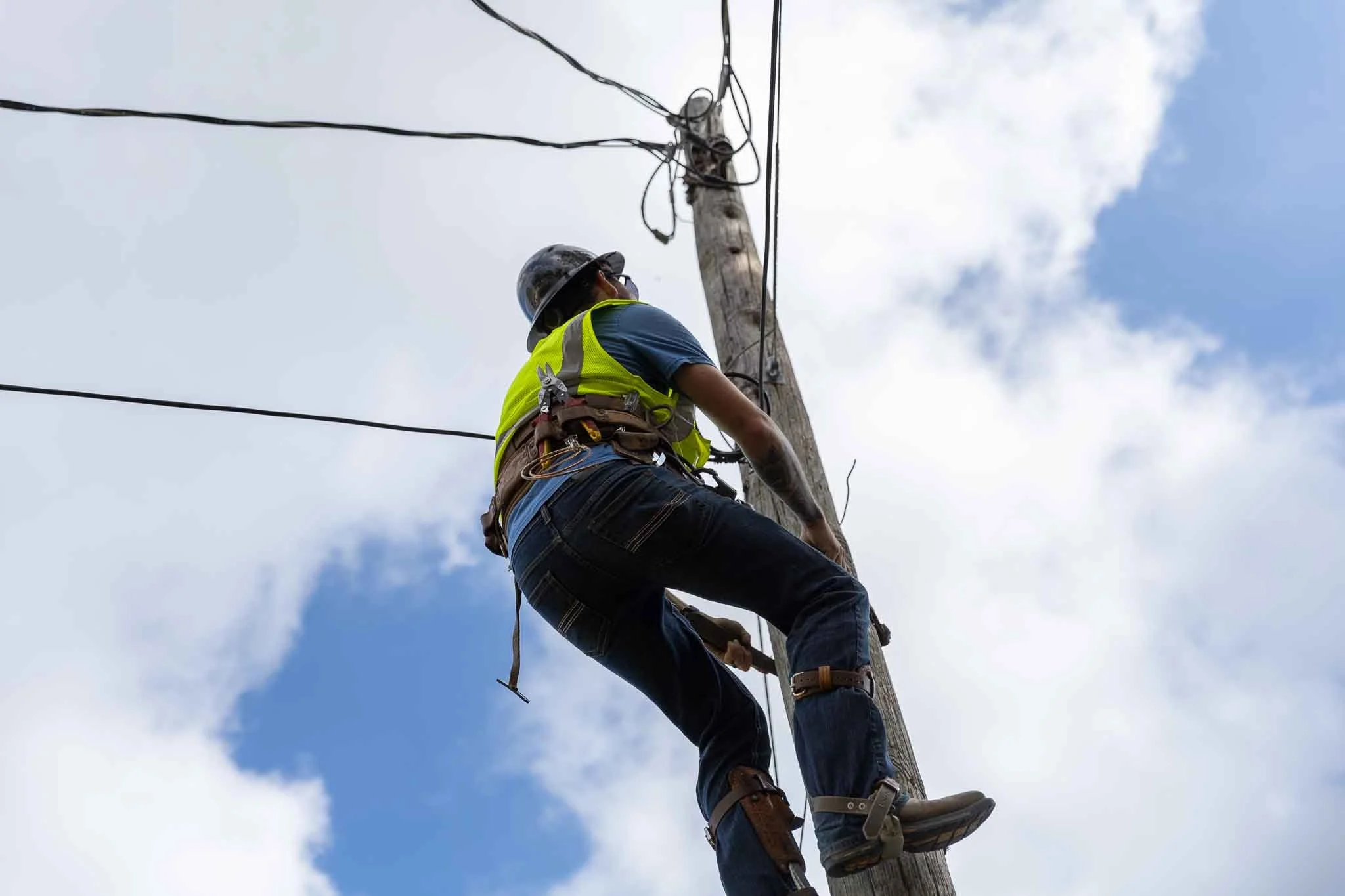 Line worker wearing a safety vest, helmet, and harness climbing a utility pole against a partly cloudy sky.
