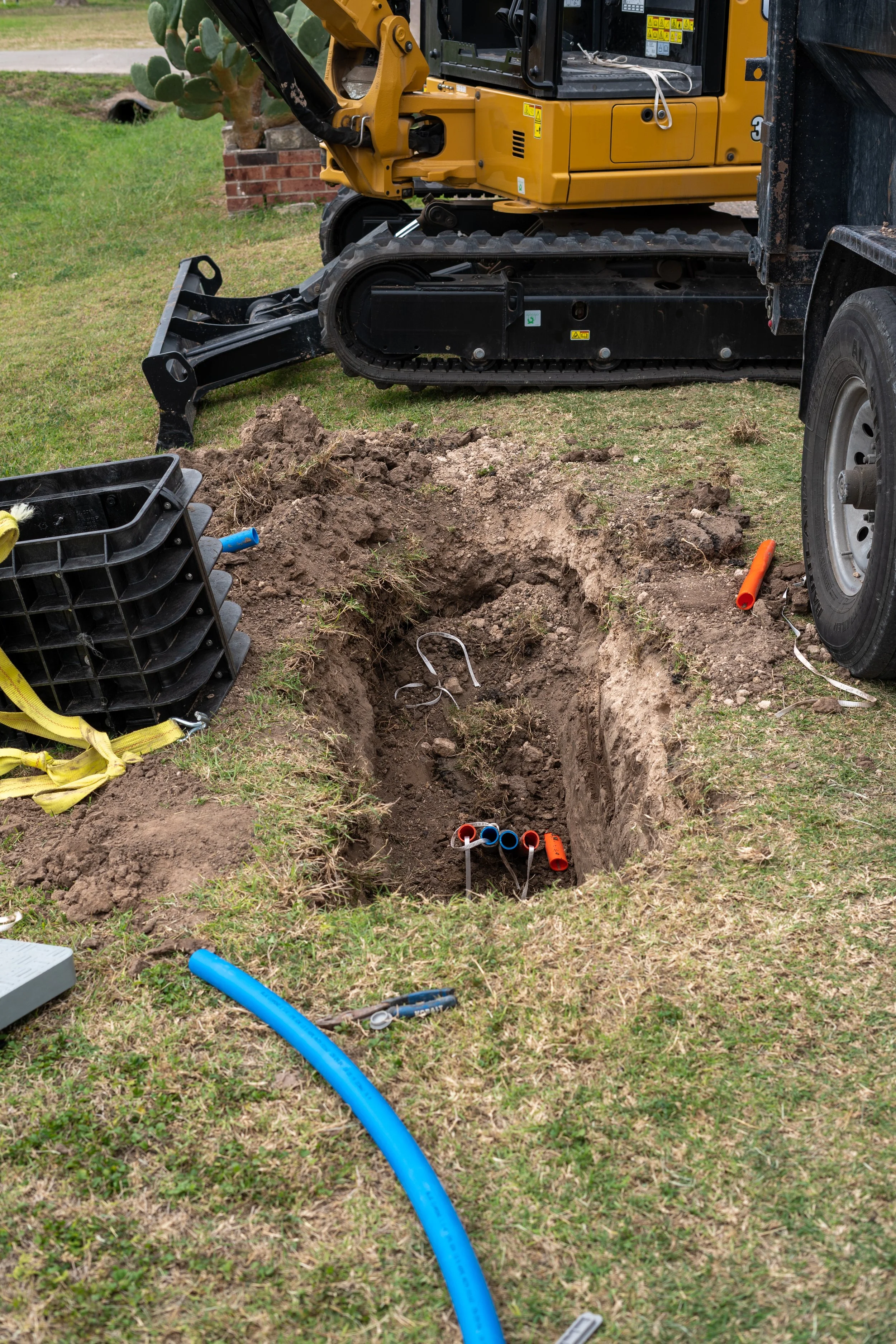 A construction site showing a deep trench in the ground with multiple colored pipes and wires inside, surrounded by earth and grass. A yellow mini excavator and a black truck are nearby with construction tools and equipment scattered around.