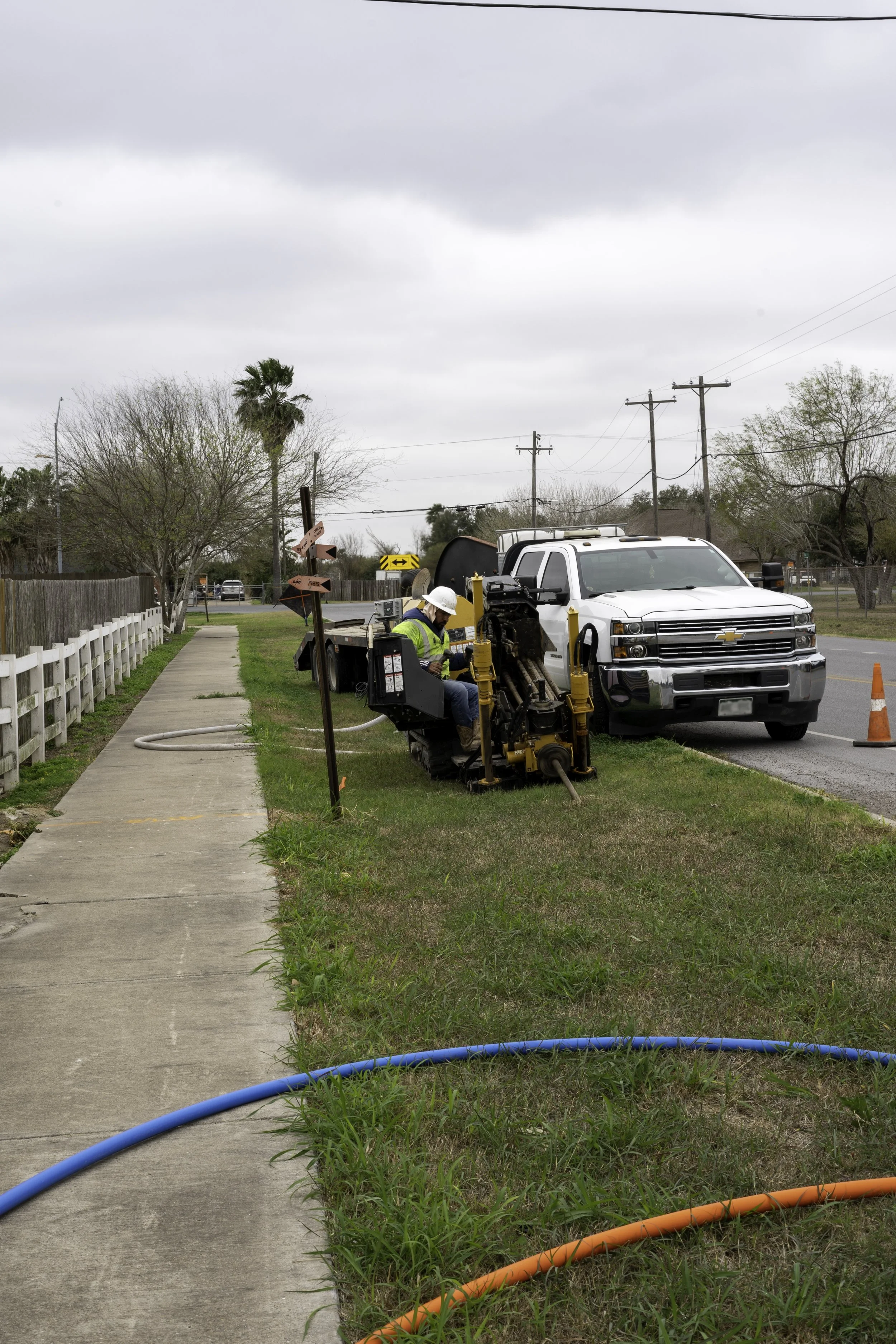 A worker in a white helmet and reflective vest operating machinery on grass next to a white pickup truck with a traffic cone on the road.