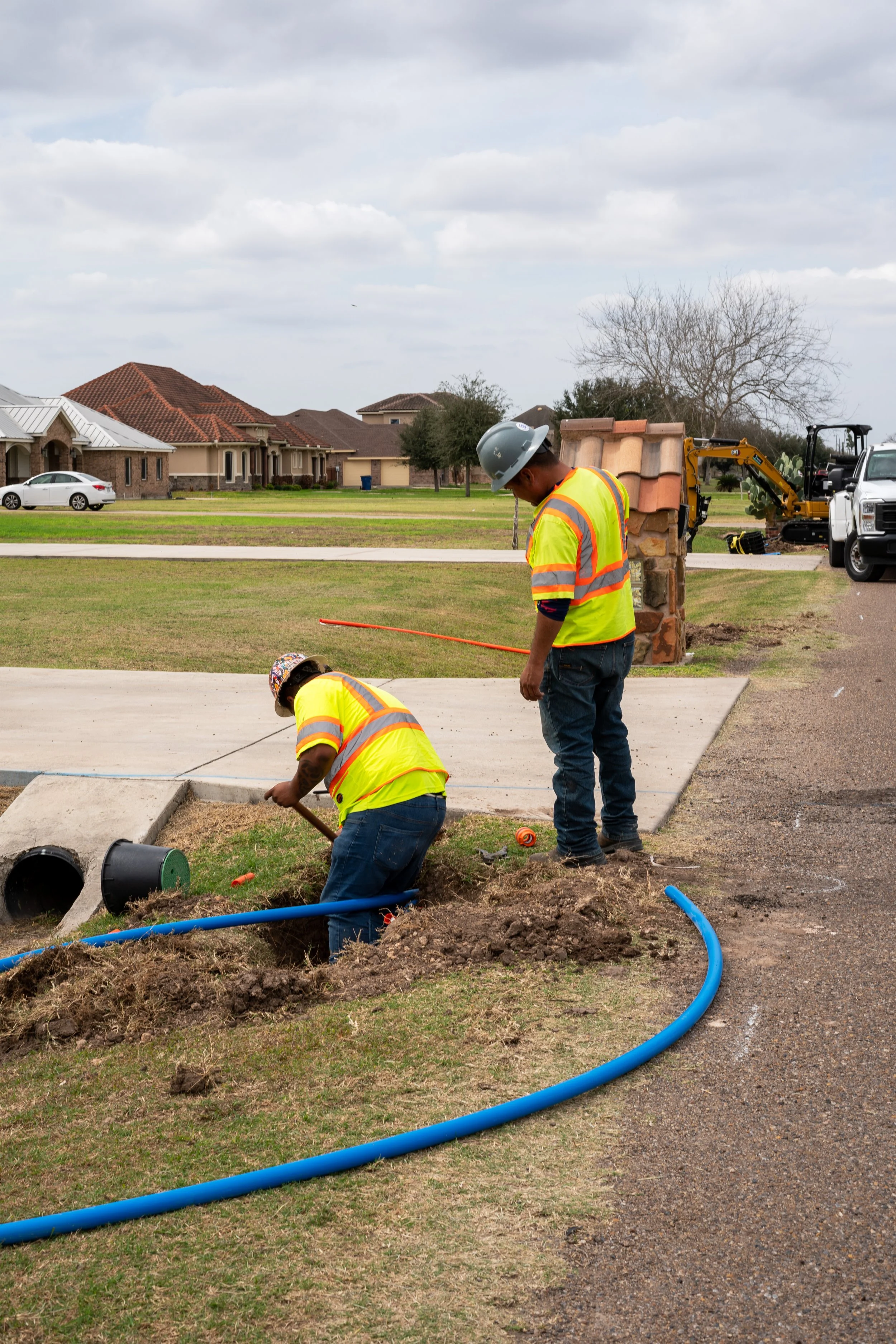 Two construction workers in safety vests and helmets working on underground piping near a residential street.