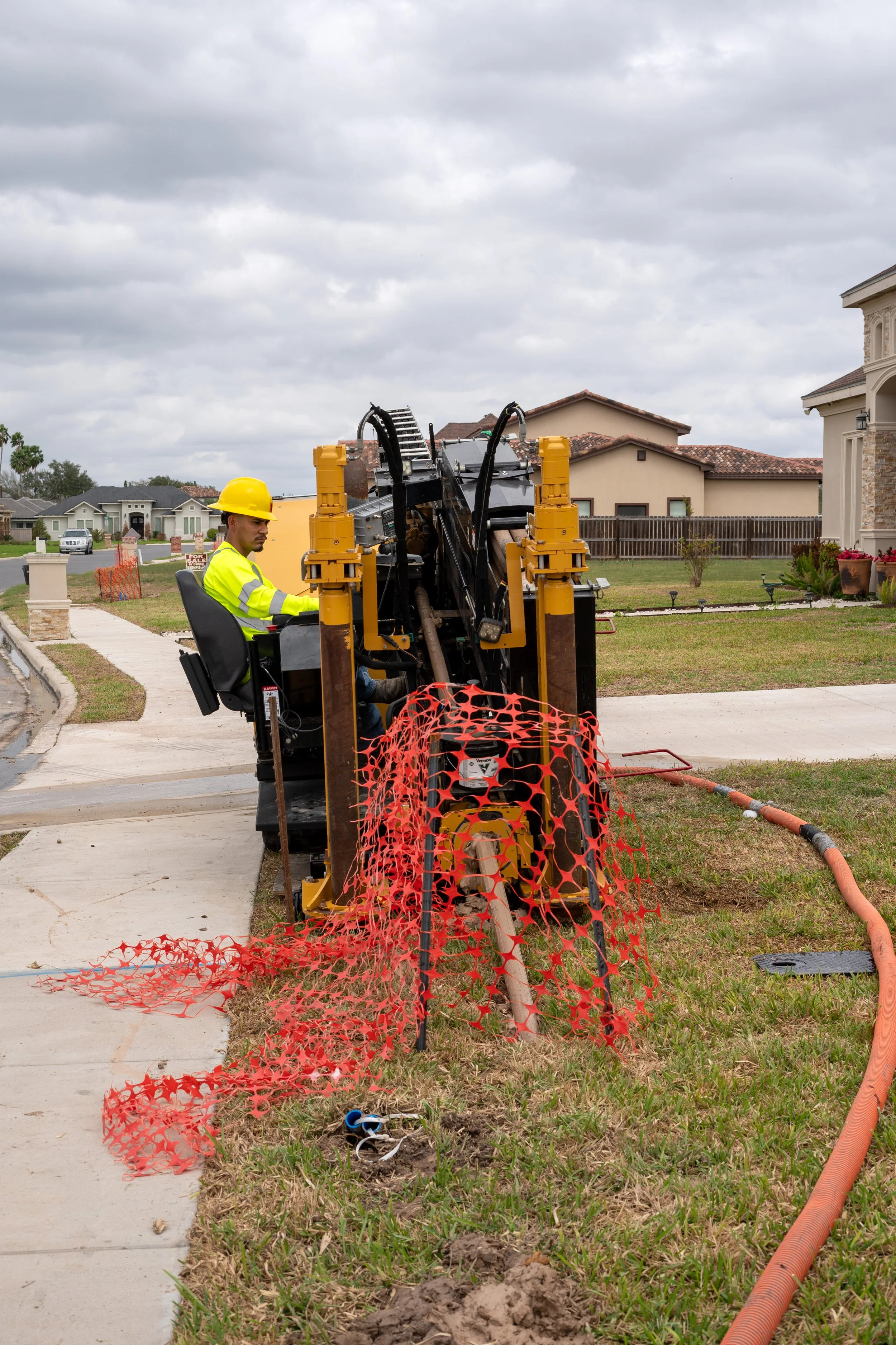 A construction worker in a yellow safety helmet and reflective vest operating machinery on a residential sidewalk, with orange safety fencing and a nearby orange pipe.