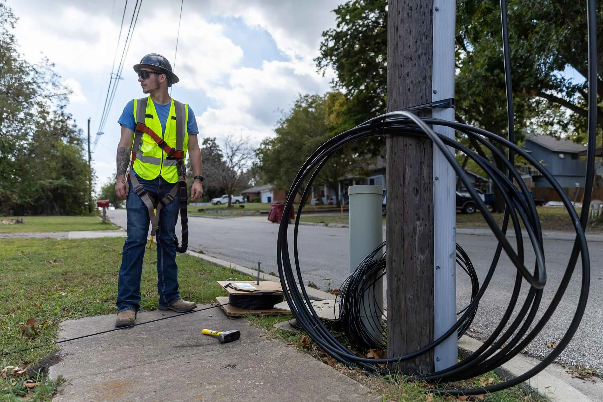 A utility worker wearing a safety helmet, reflective vest, and harness working on power lines outside on a residential street.