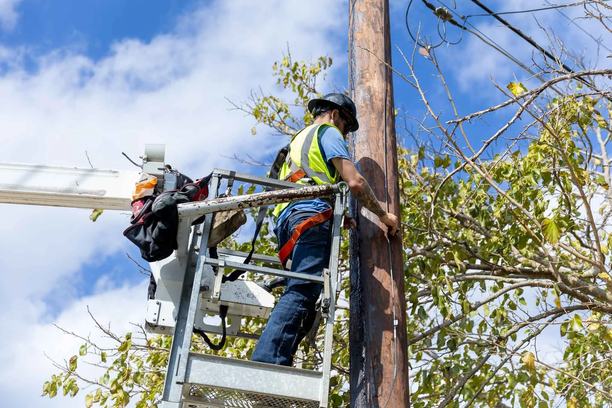 A worker in a safety vest and helmet on a lift pole working on a power line during daytime.