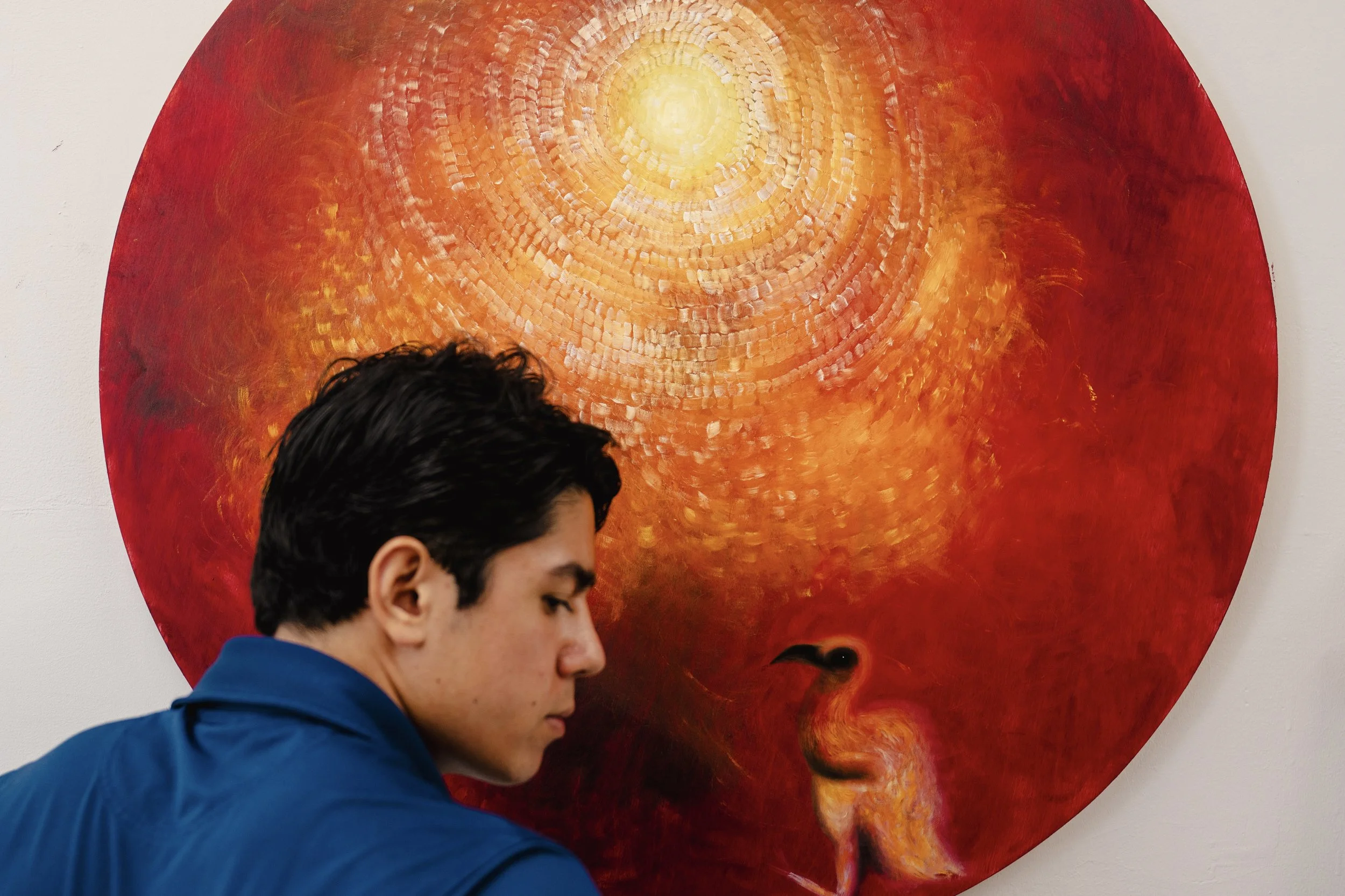 A young man with dark curly hair wearing a blue jacket, looking downward in front of a large, circular, yellow and orange abstract painting on a white wall.