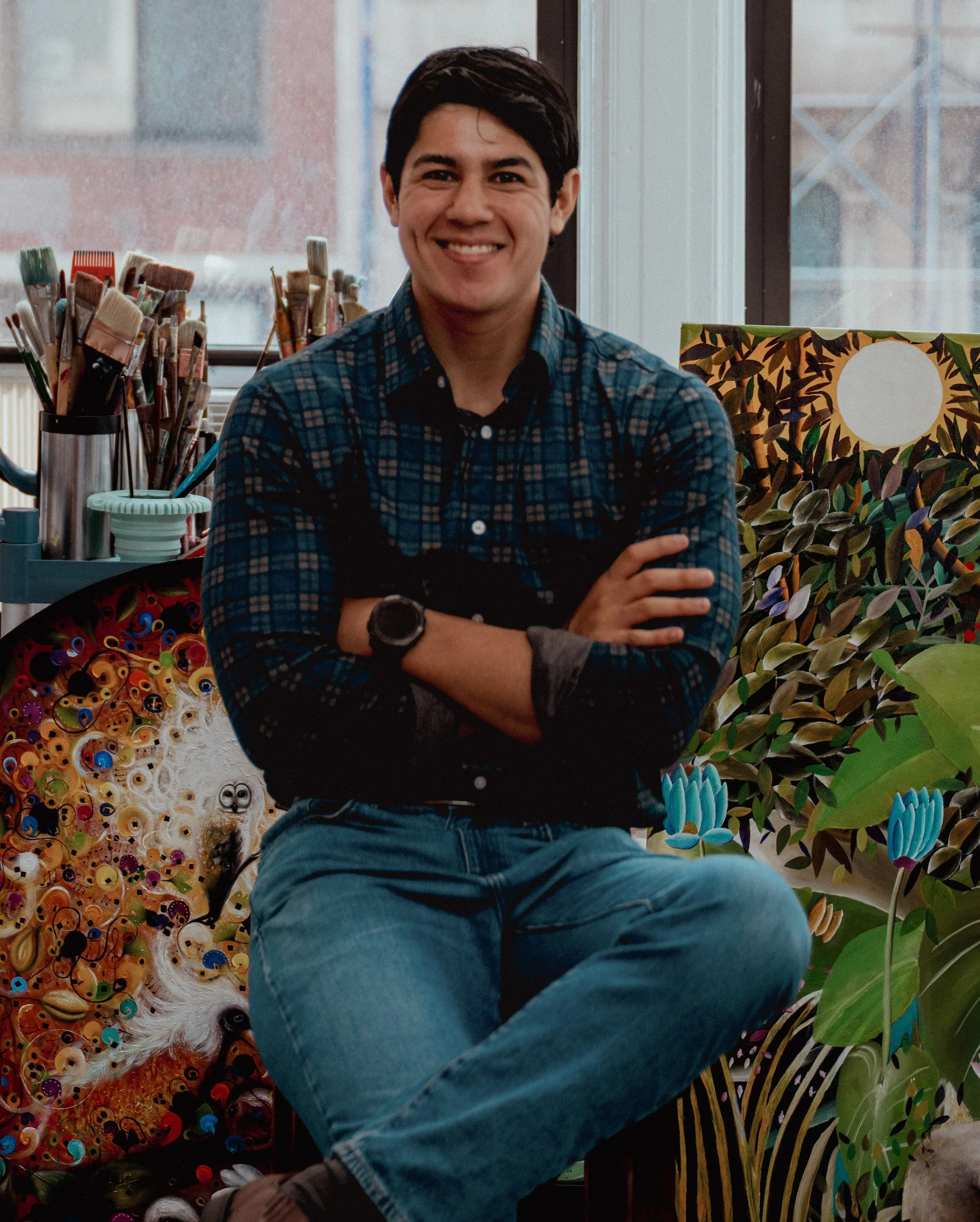A young man with dark hair, wearing a plaid shirt, sitting cross-legged with arms crossed, smiling in an art studio with colorful paintings and art supplies around him.