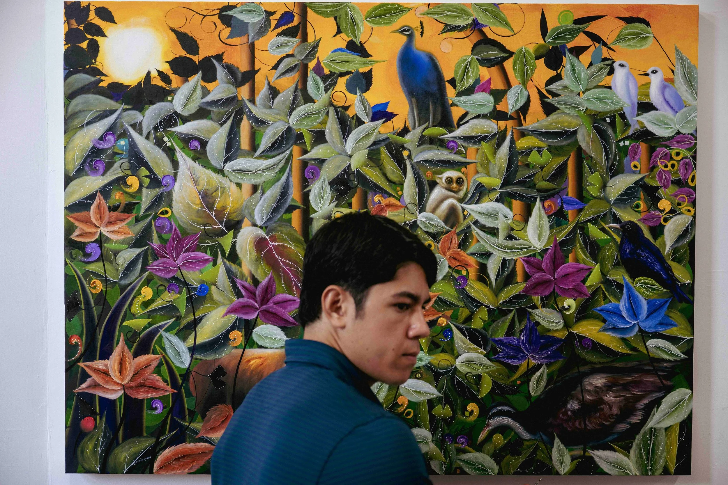Brightly colored abstract painting of a jungle scene with leaves, flowers, and birds, including a blue bird, white birds, and a black bird, set against a sunset or sunrise background. A person is partially visible in the foreground.