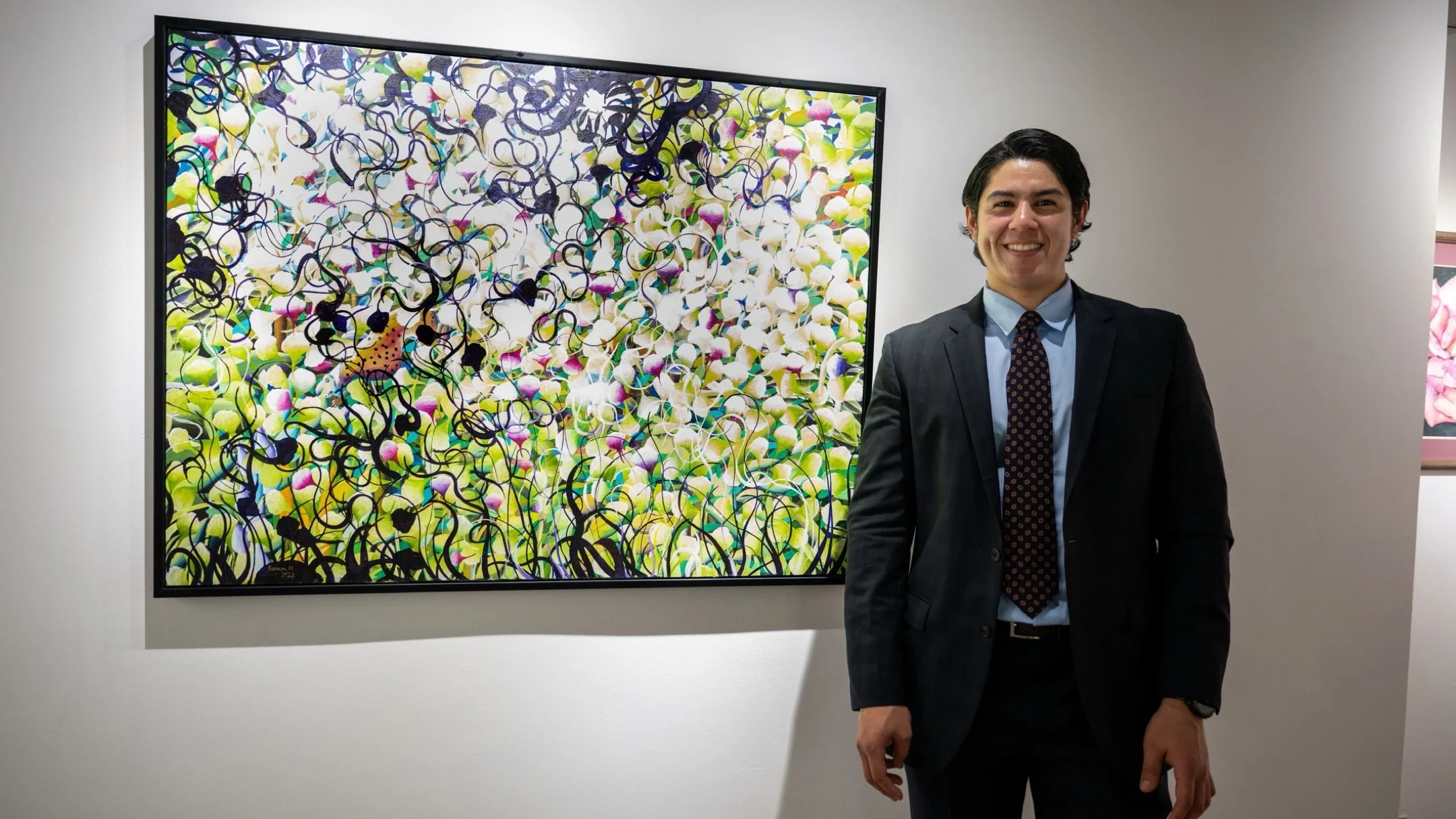 A man in a dark suit and tie standing next to a colorful abstract painting at an art gallery.