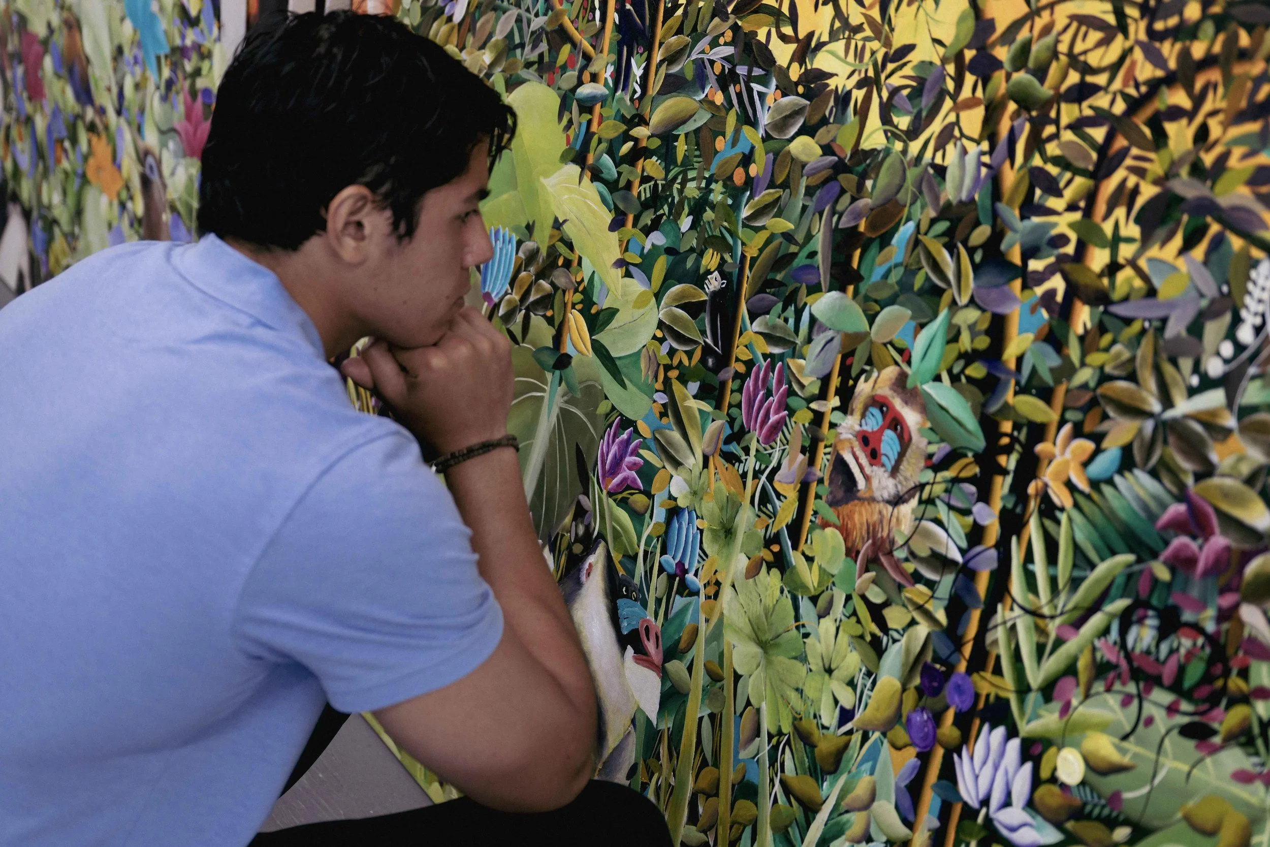 A person with dark hair and a blue shirt is leaning forward and observing in front of a colorful, intricate wall of layered, painted foliage and flowers with a butterfly and butterfly mask near the bottom.