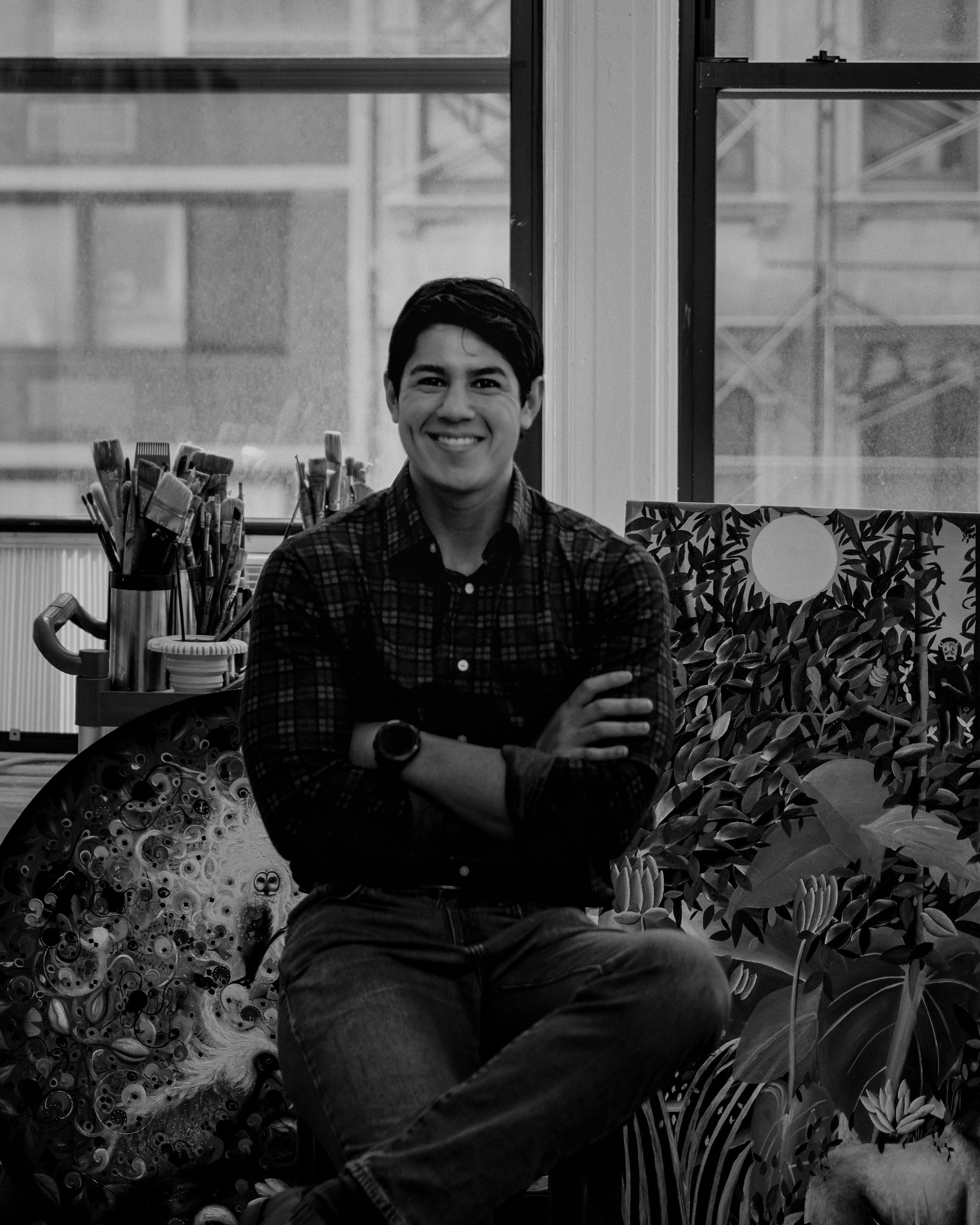 A smiling young man wearing a plaid shirt and watch, sitting with crossed arms in a creative studio with art supplies, plants, and artwork.