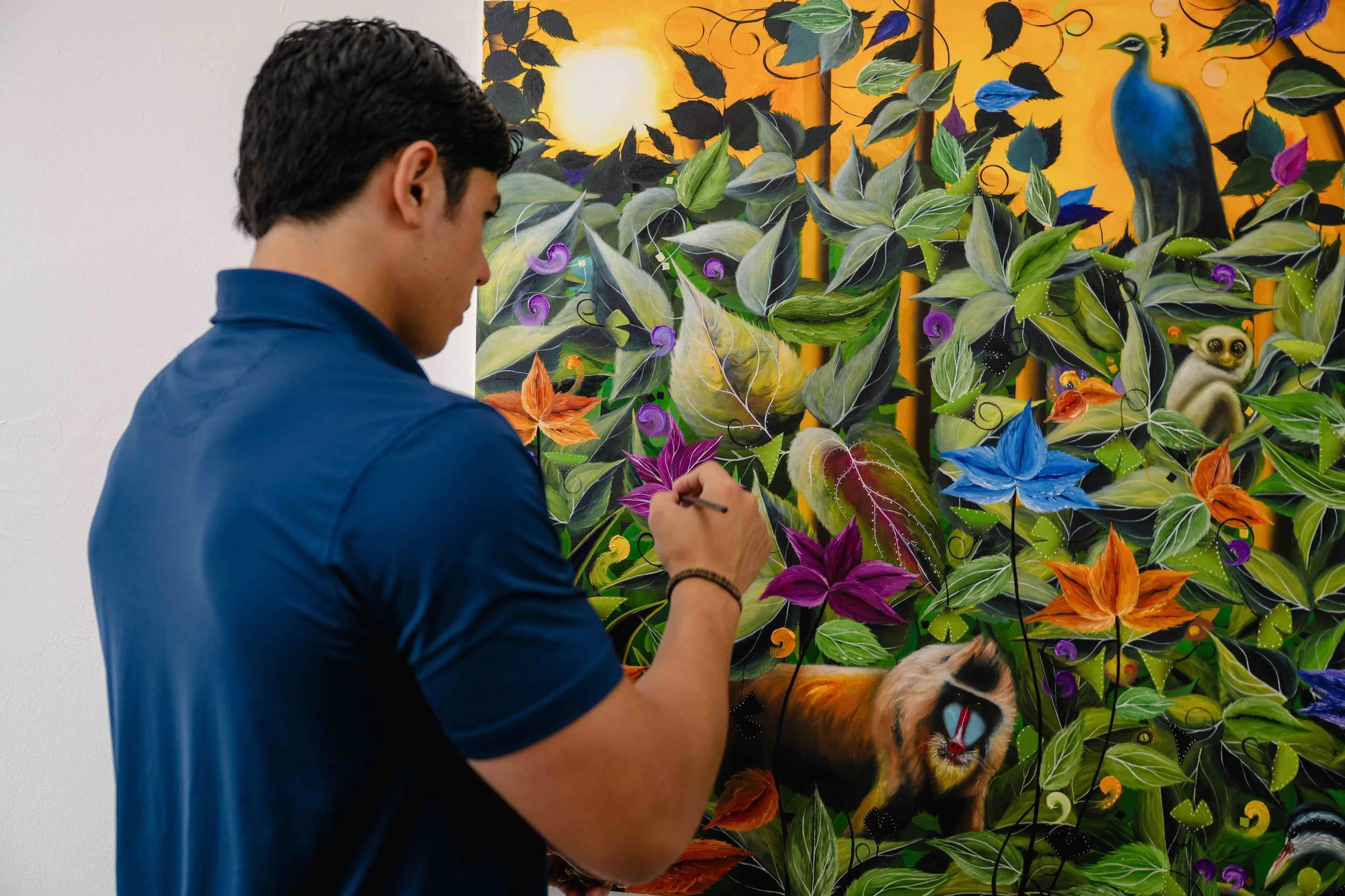 A man painting a colorful and intricate artwork featuring leaves, flowers, a peacock, a lion, a lemur, and abstract shapes on a large canvas in an art gallery.