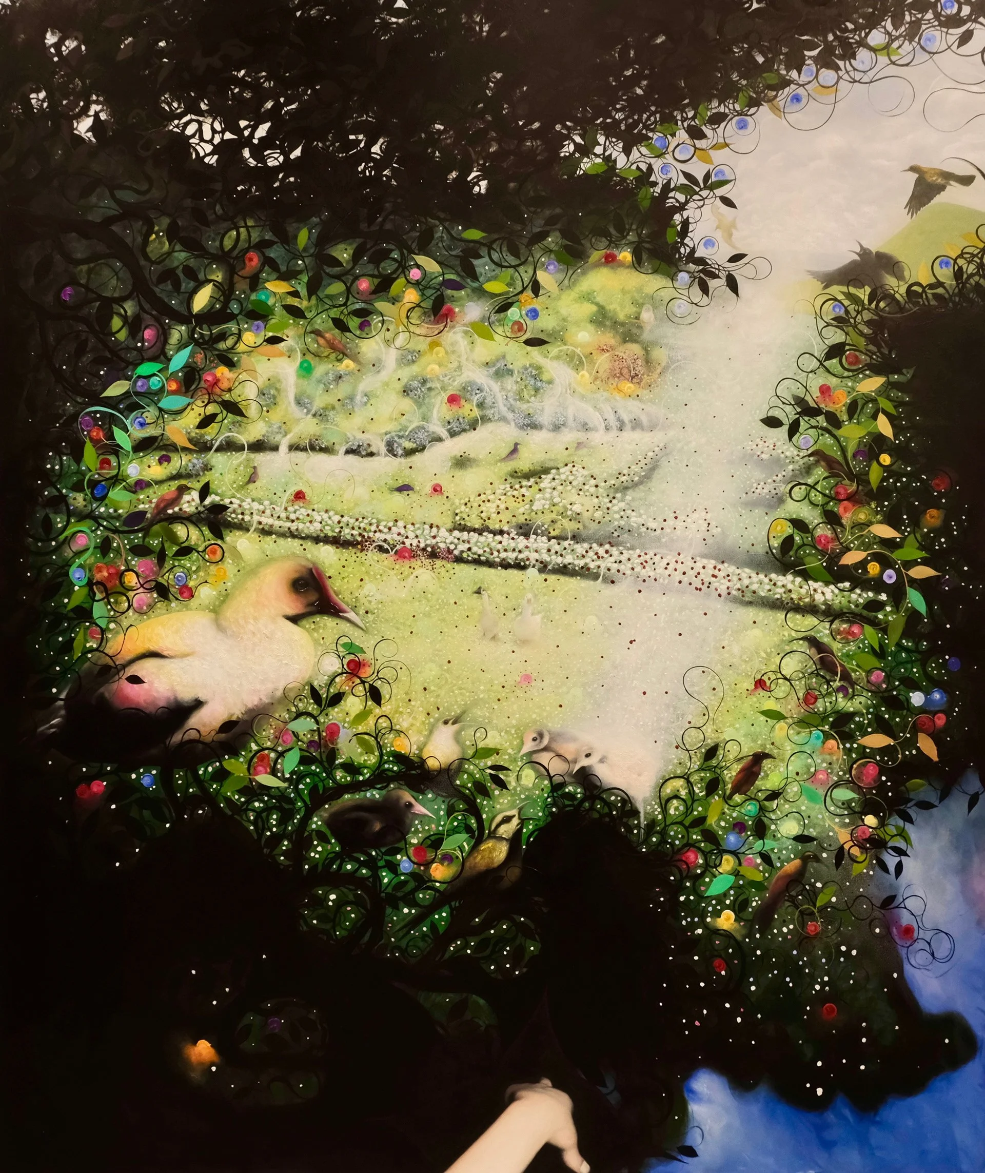 A person holds a white cat by the tail while other cats sit on a lush grassy area under vibrant, swirling foliage with colorful orbs and several birds perched and flying nearby.
