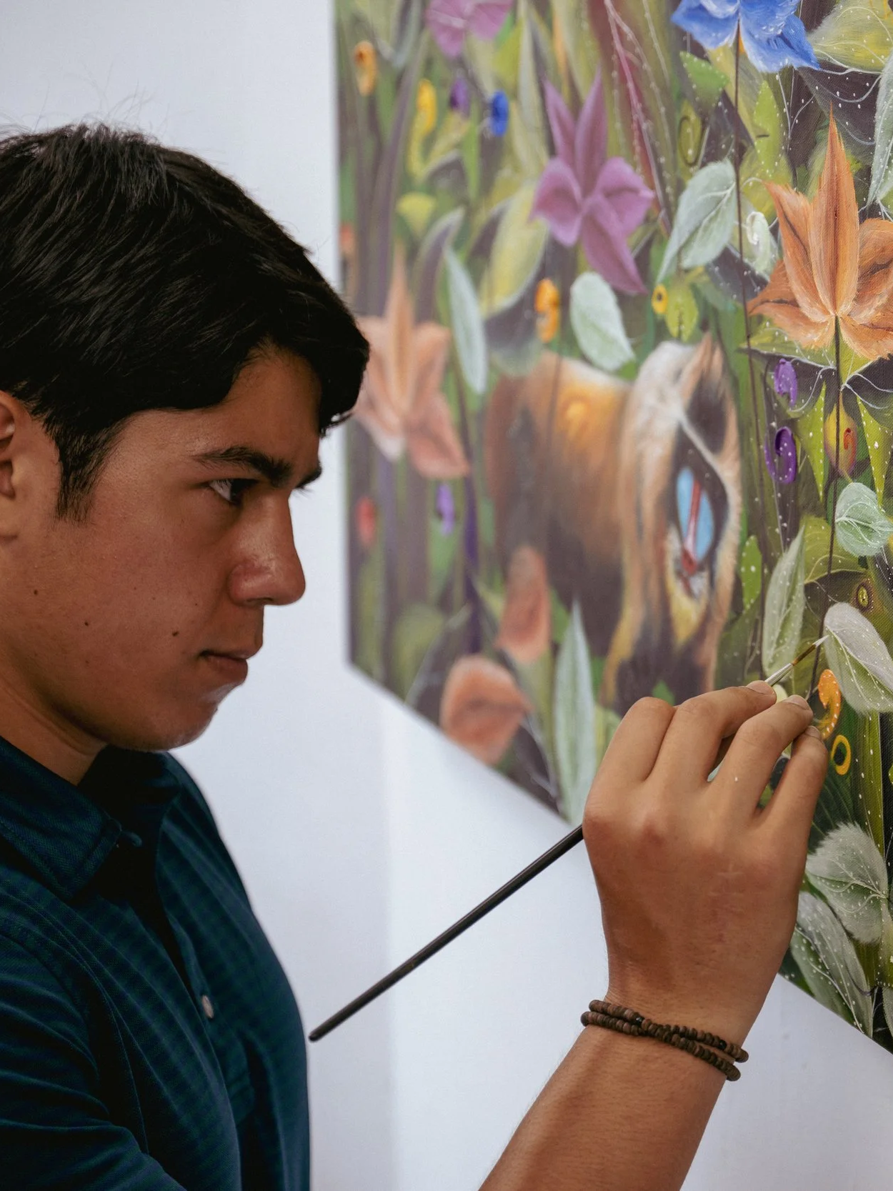 A young man with dark hair carefully restoring or painting a colorful, detailed artwork of a jungle scene with plants and animals.