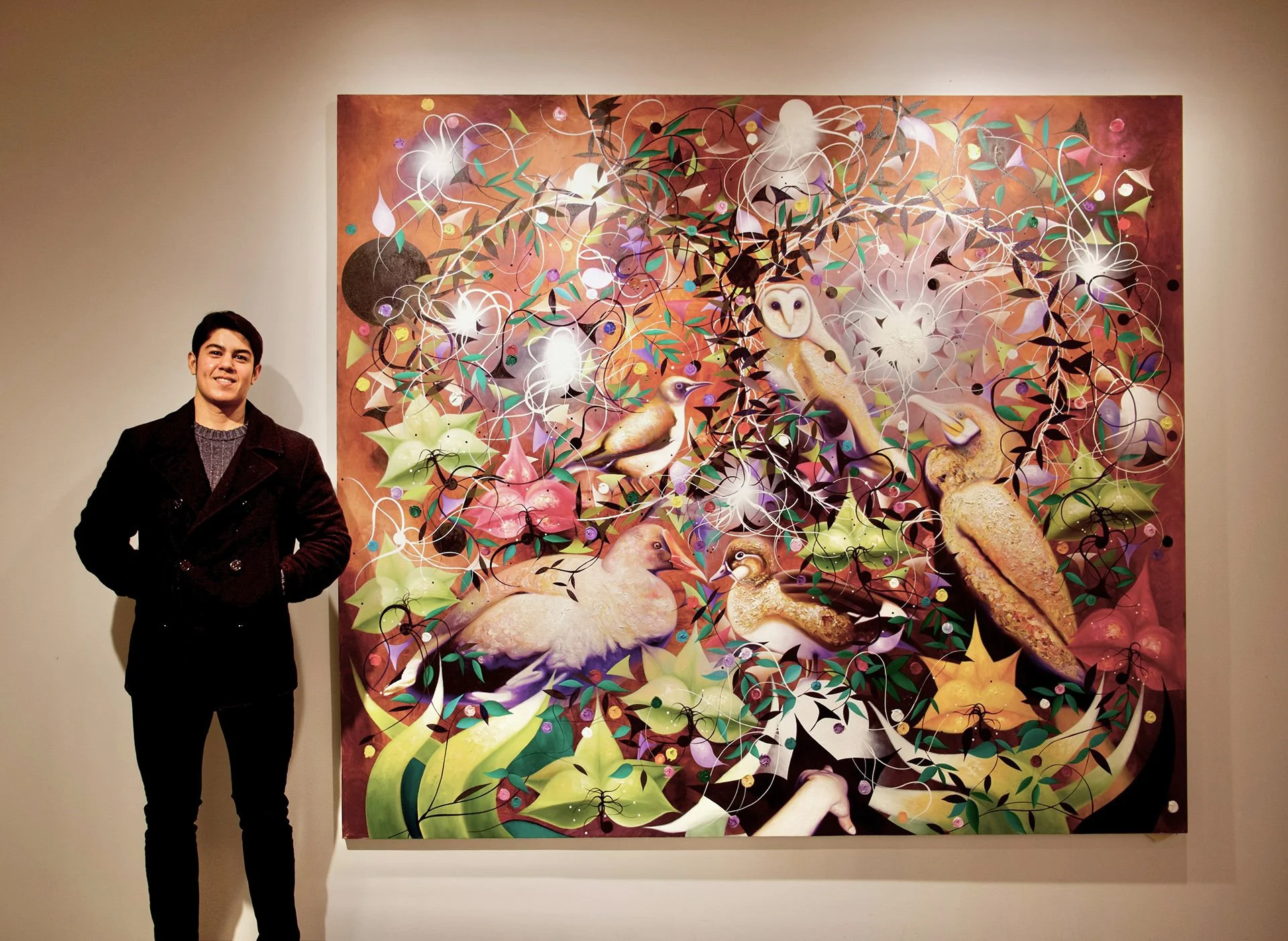 A young man standing next to a colorful abstract painting with birds, leaves, and swirling patterns in an art gallery.