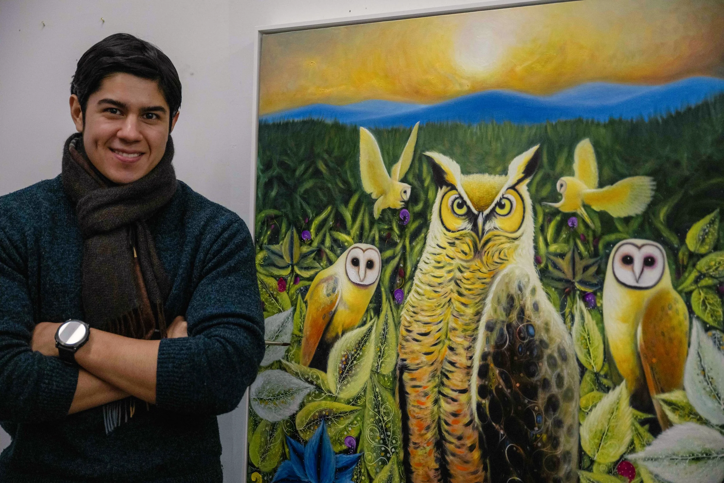 A young man standing next to a colorful painting of owls and birds in a lush green field during sunset.