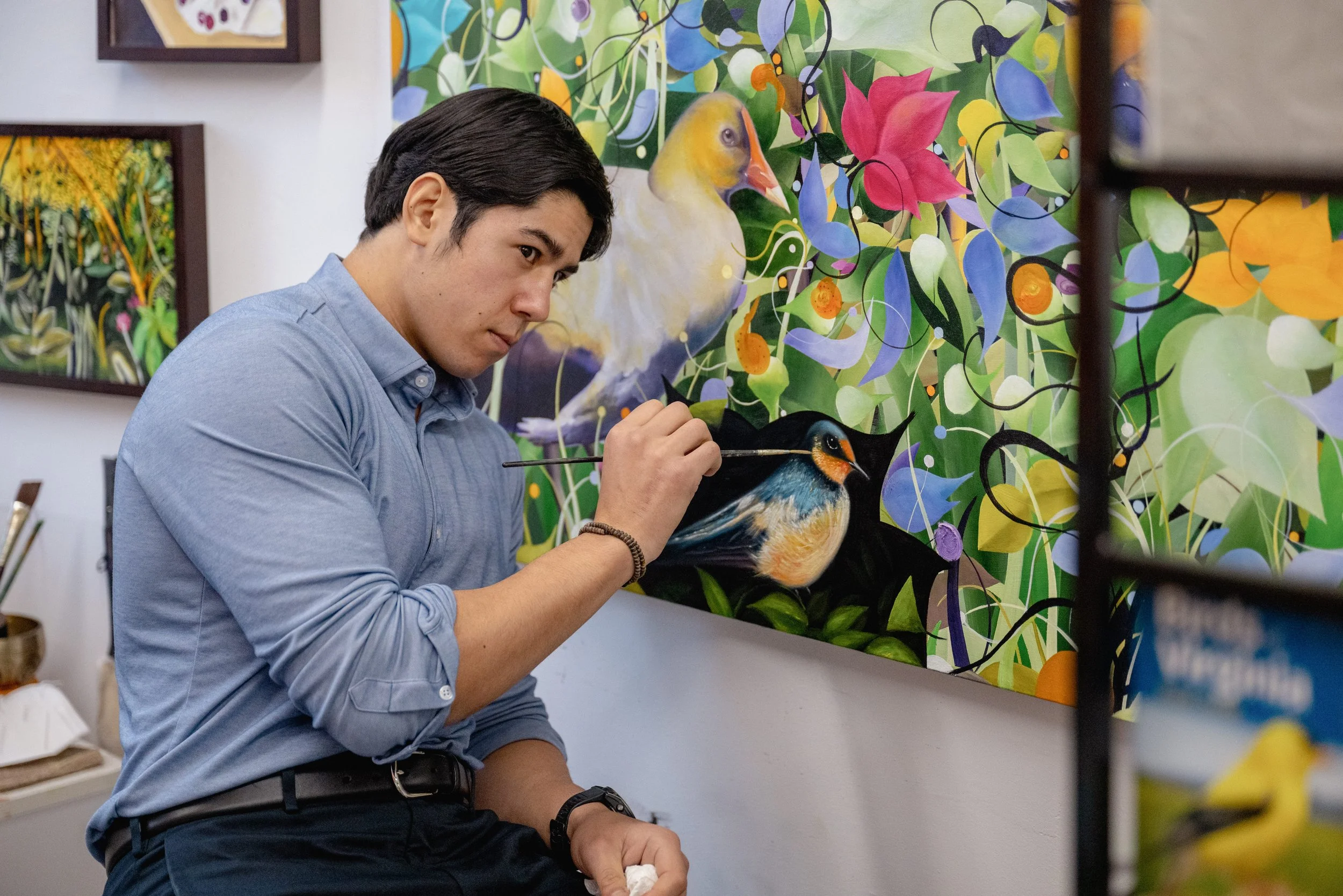 A man in a blue shirt painting a bird on a canvas in an art gallery decorated with colorful bird and floral paintings.