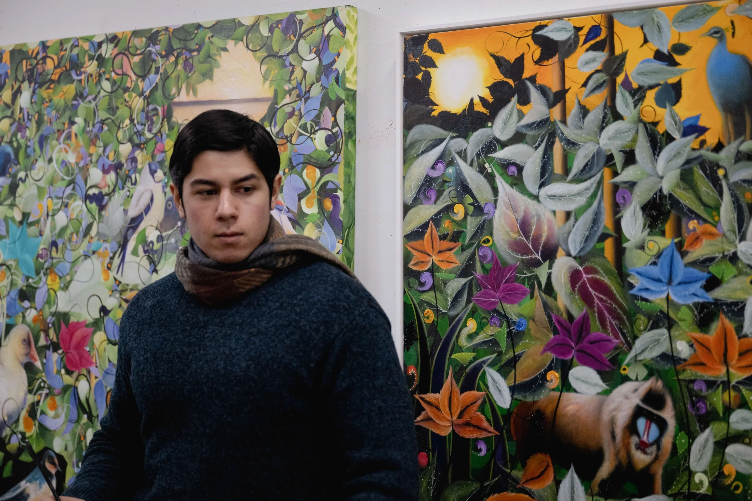 A young man with dark hair, wearing a dark sweater and scarf, standing in front of colorful botanical paintings in an art gallery.