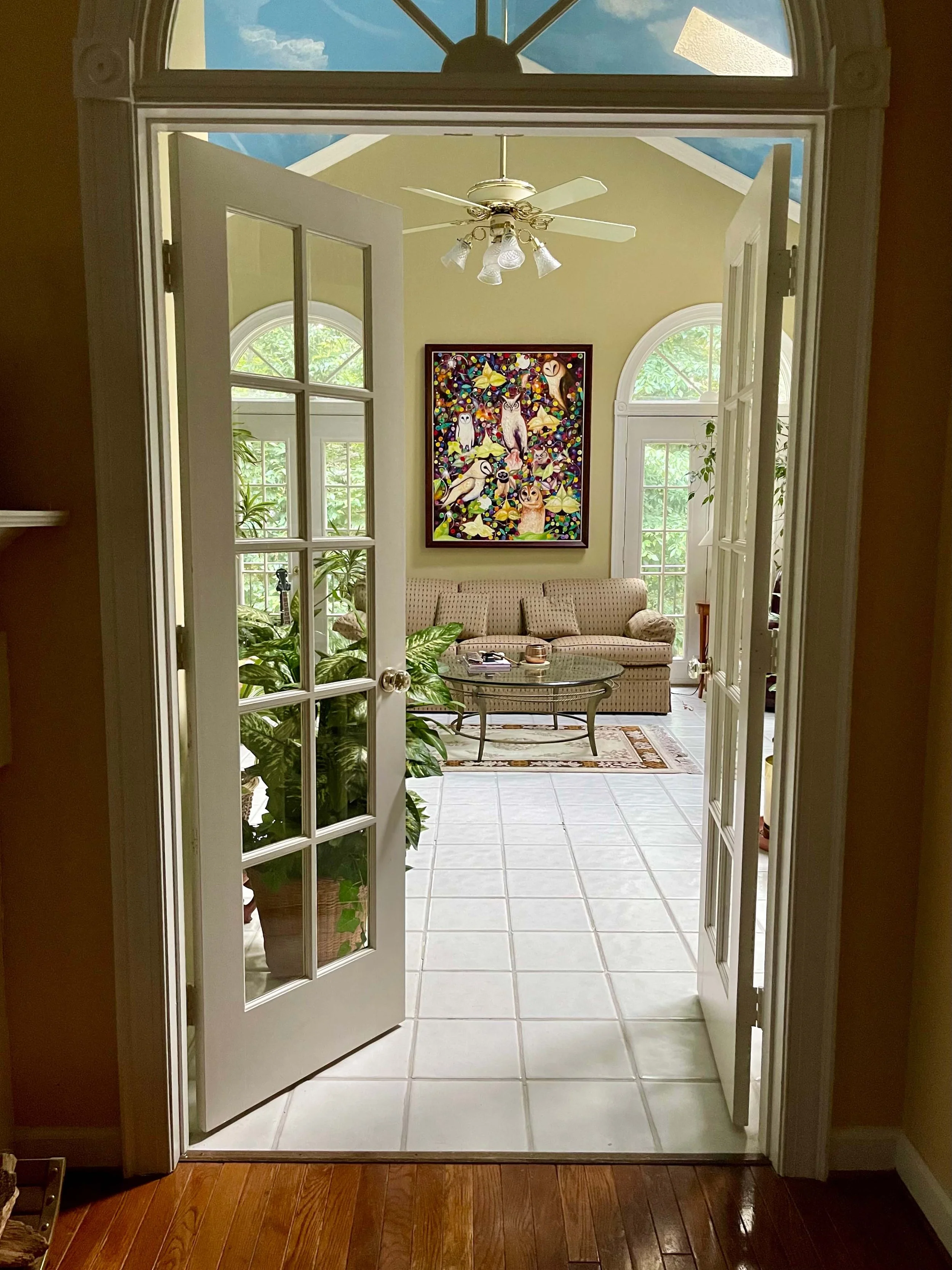 Looking through a doorway into a sunlit living room with large arched windows, a ceiling fan, a colorful owl-themed painting, a beige sofa, and various plants.