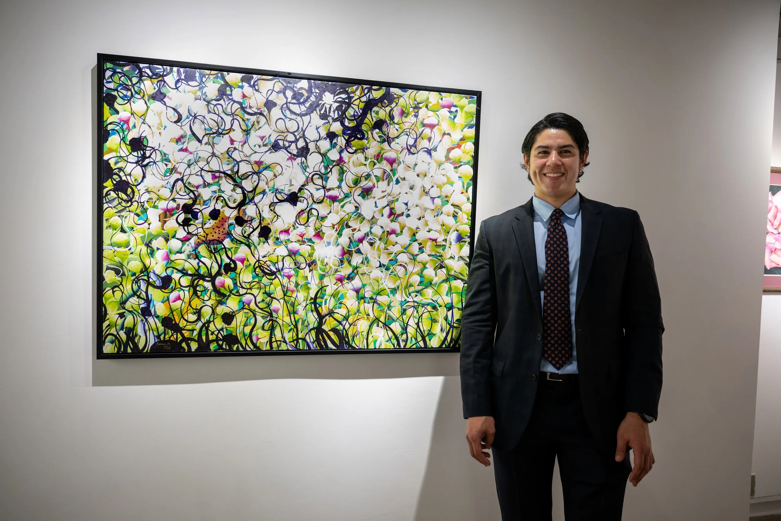 A man in a dark suit, light blue shirt, and patterned tie standing next to a colorful abstract painting in an art gallery.