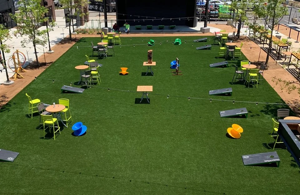 Outdoor seating area with tables, chairs, and bean bag chairs on artificial grass, surrounded by trees and a black fence, with a stage in the background.