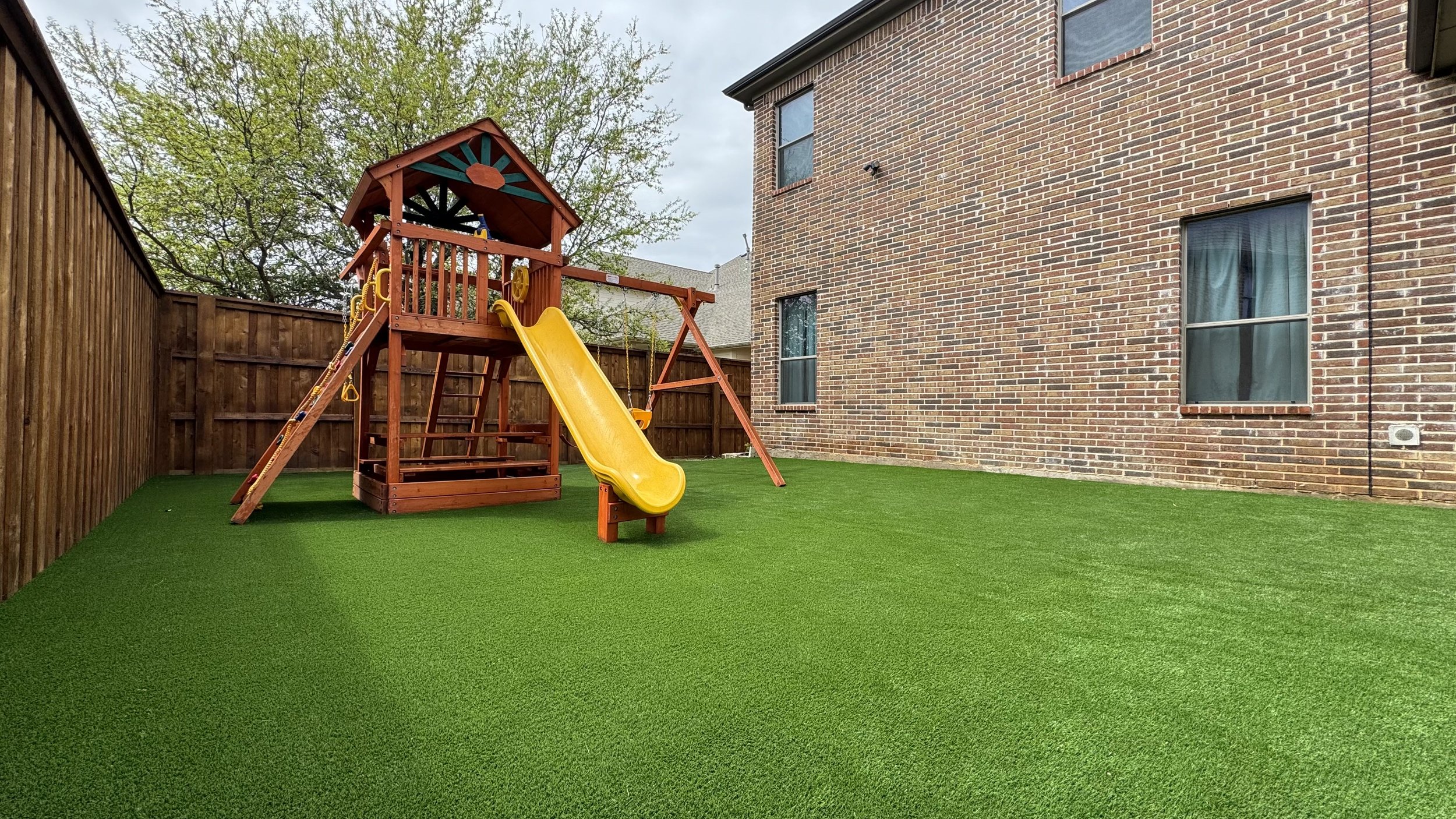 A small backyard with artificial green grass, enclosed by a wooden fence. There is a wooden playset with a yellow slide near a brick house. The playset has a roof, swings, and a ladder. A large tree with green leaves is visible in the background, and the sky is cloudy.