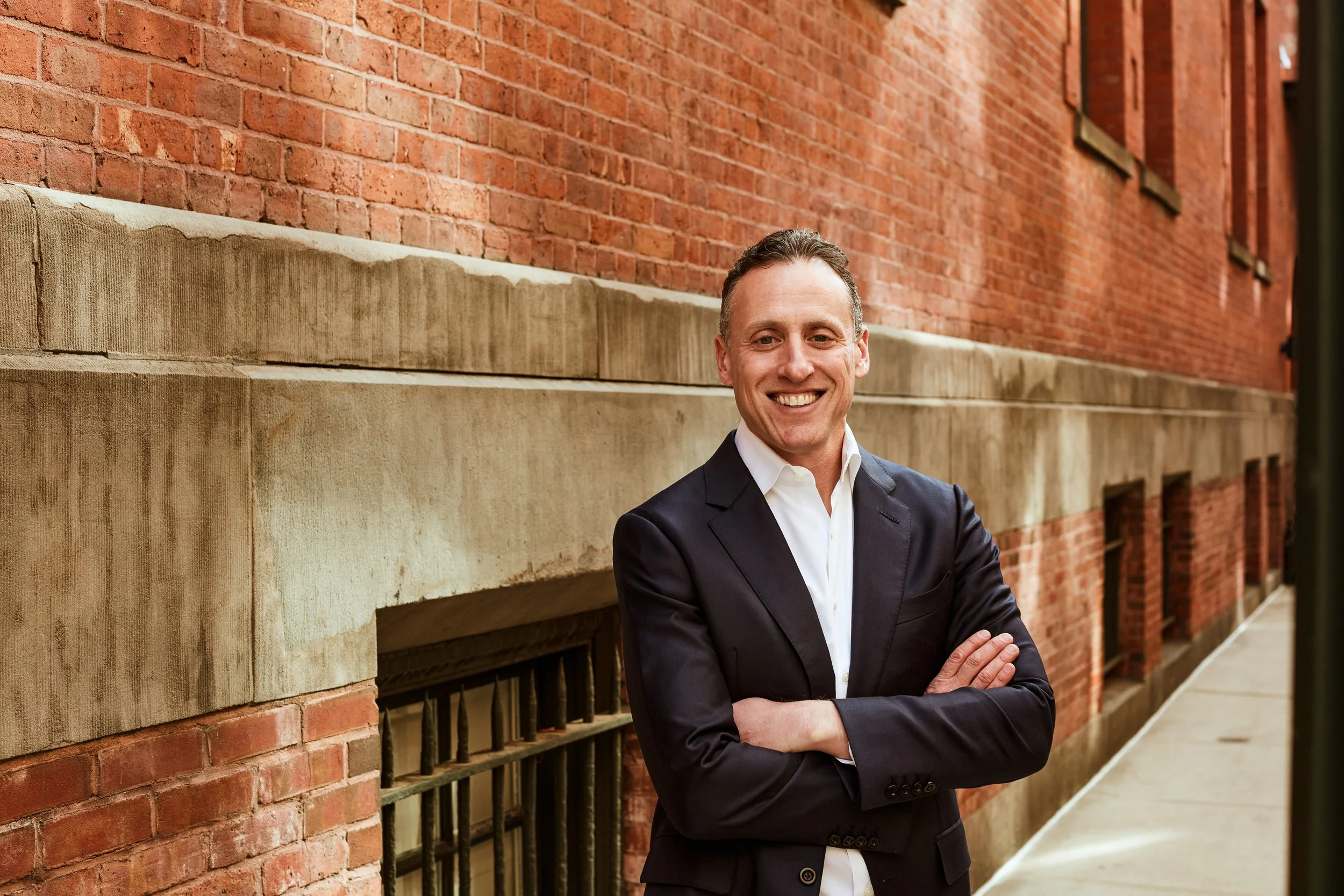 Smiling man in a business suit standing outdoors against a brick building wall with crossed arms.