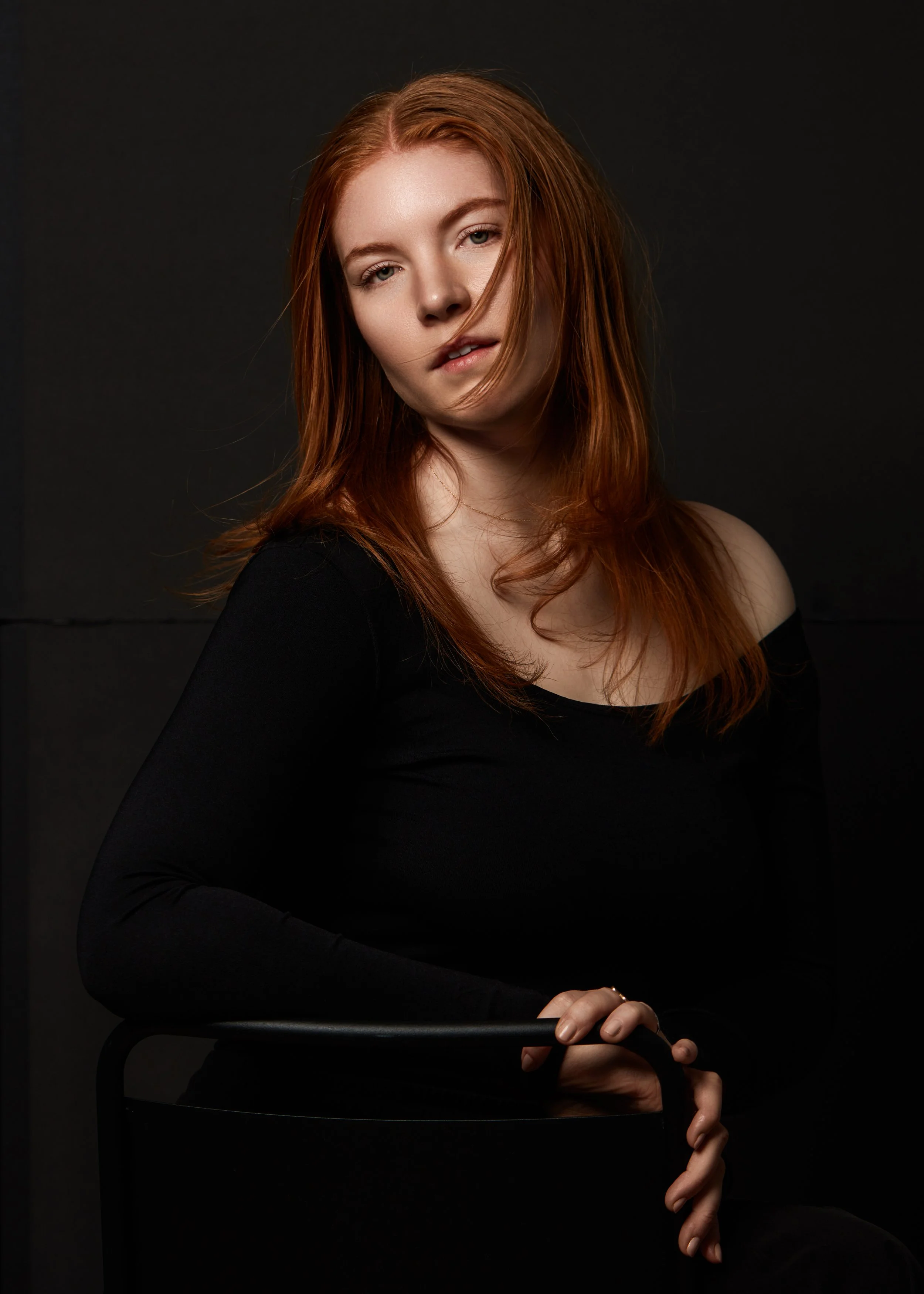 A young woman with red hair and fair skin posing against a dark backdrop, wearing a black off-the-shoulder top, with a neutral facial expression.