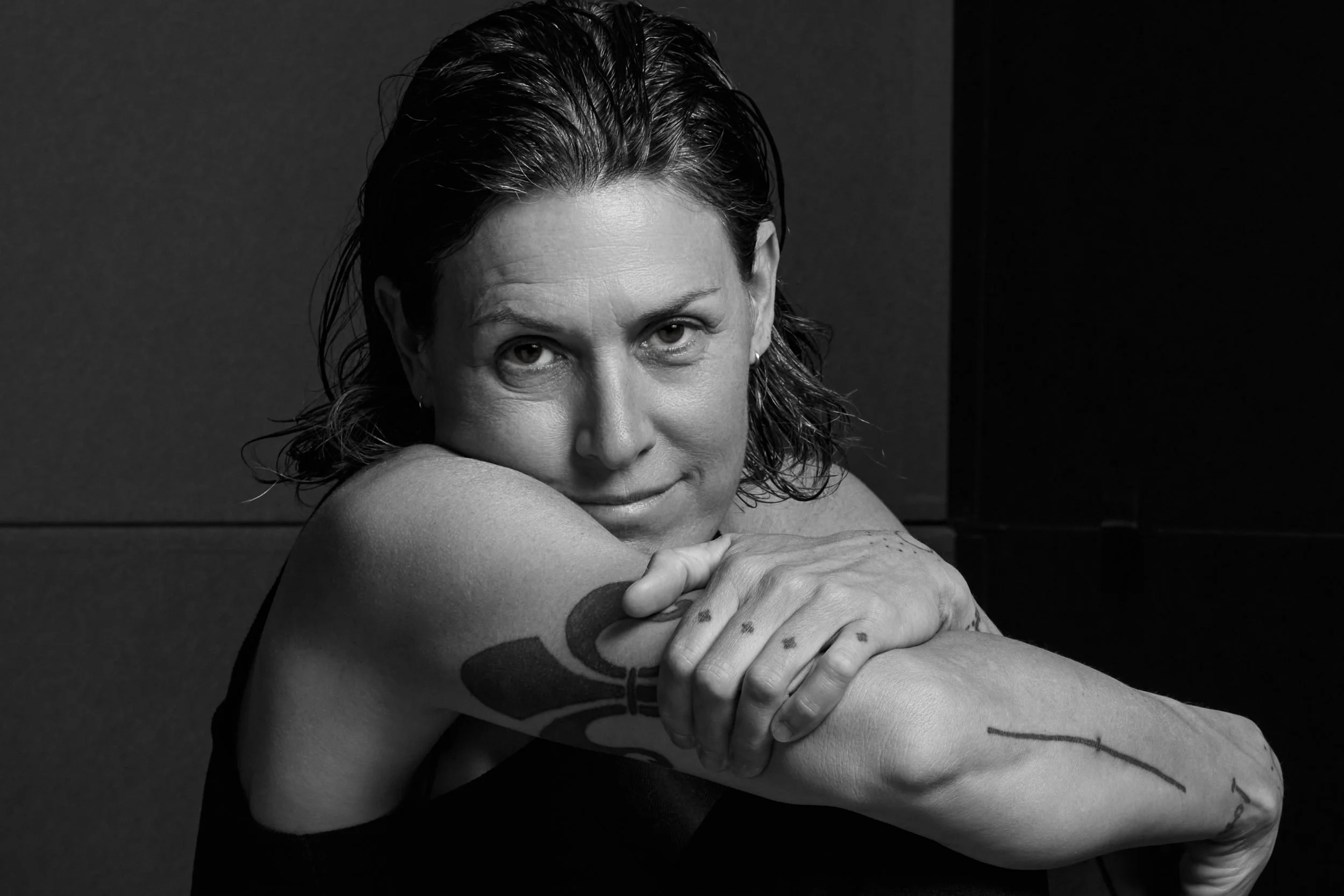 A woman with short dark hair and tattoos on her arms, resting her arms on her bent knee and smiling softly at the camera.