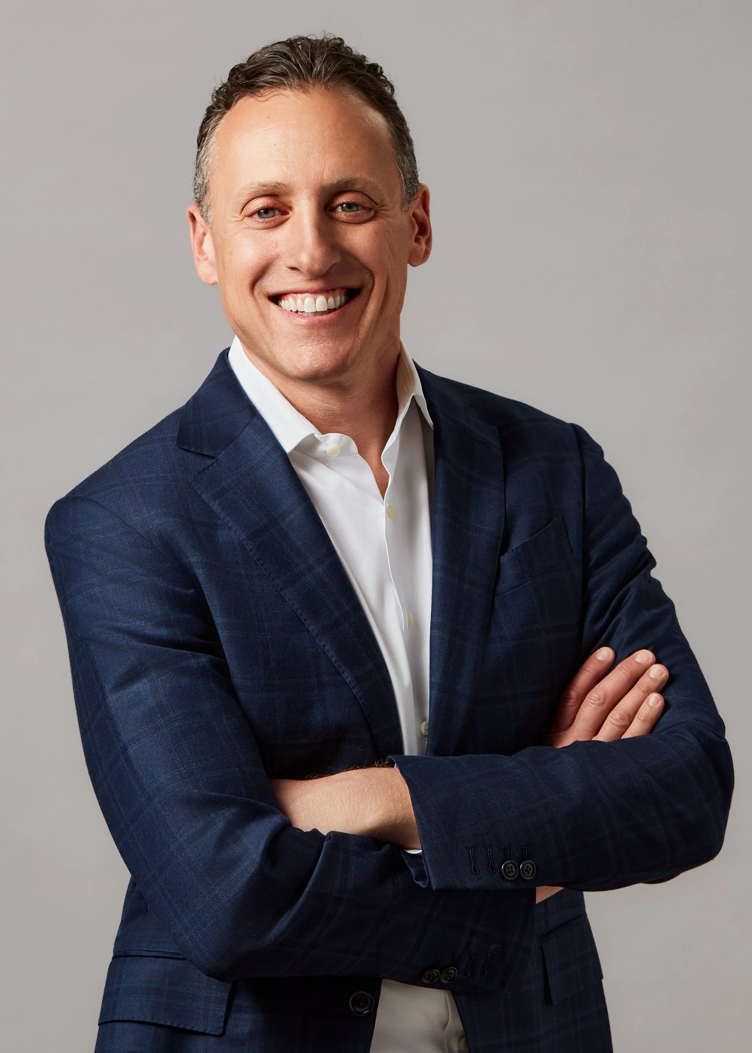 A smiling man with light skin, short light brown hair, wearing a navy blue checkered suit and white dress shirt, standing with arms crossed against a plain gray background.