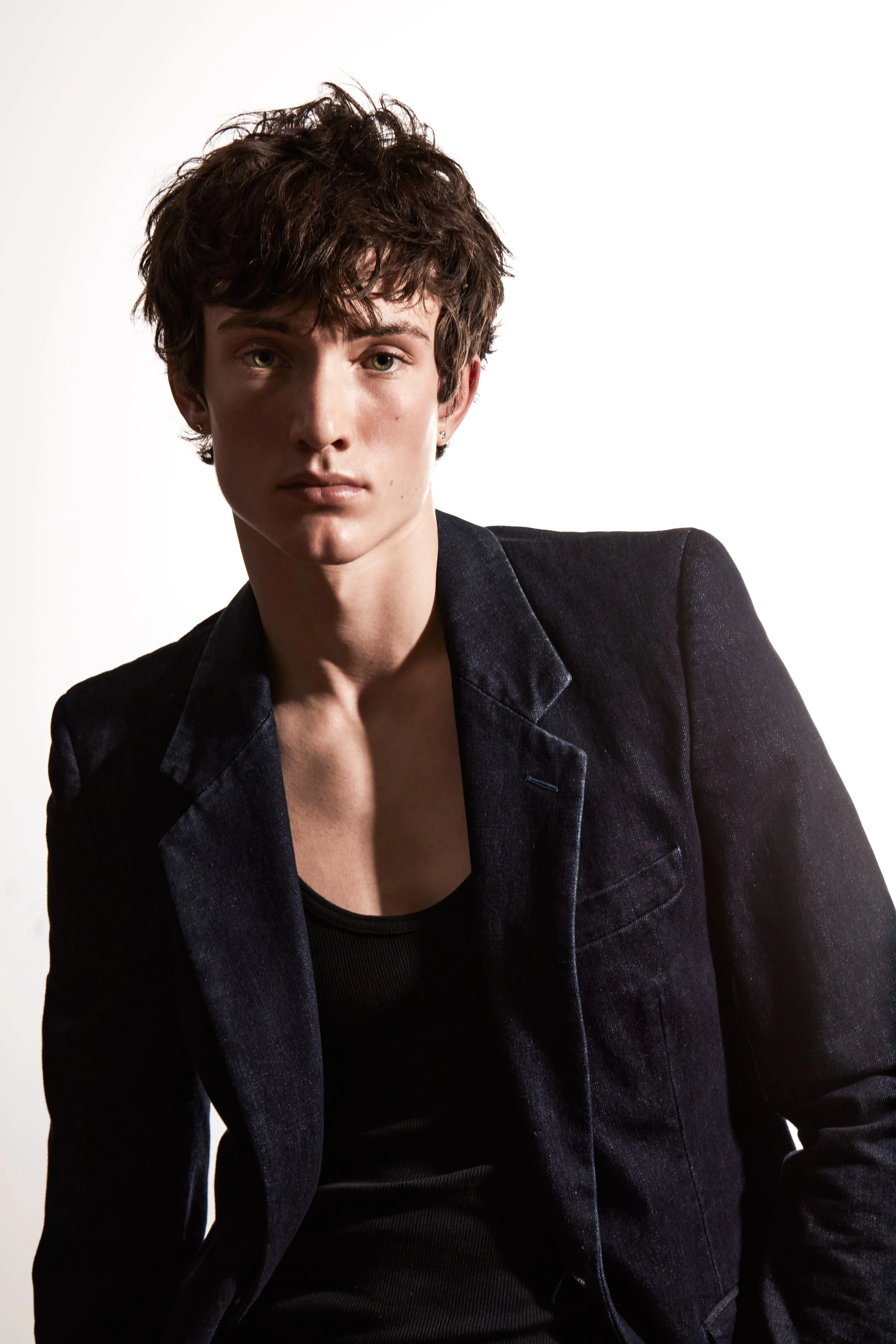 Young man with curly brown hair wearing a dark blazer and black shirt, looking directly at the camera.