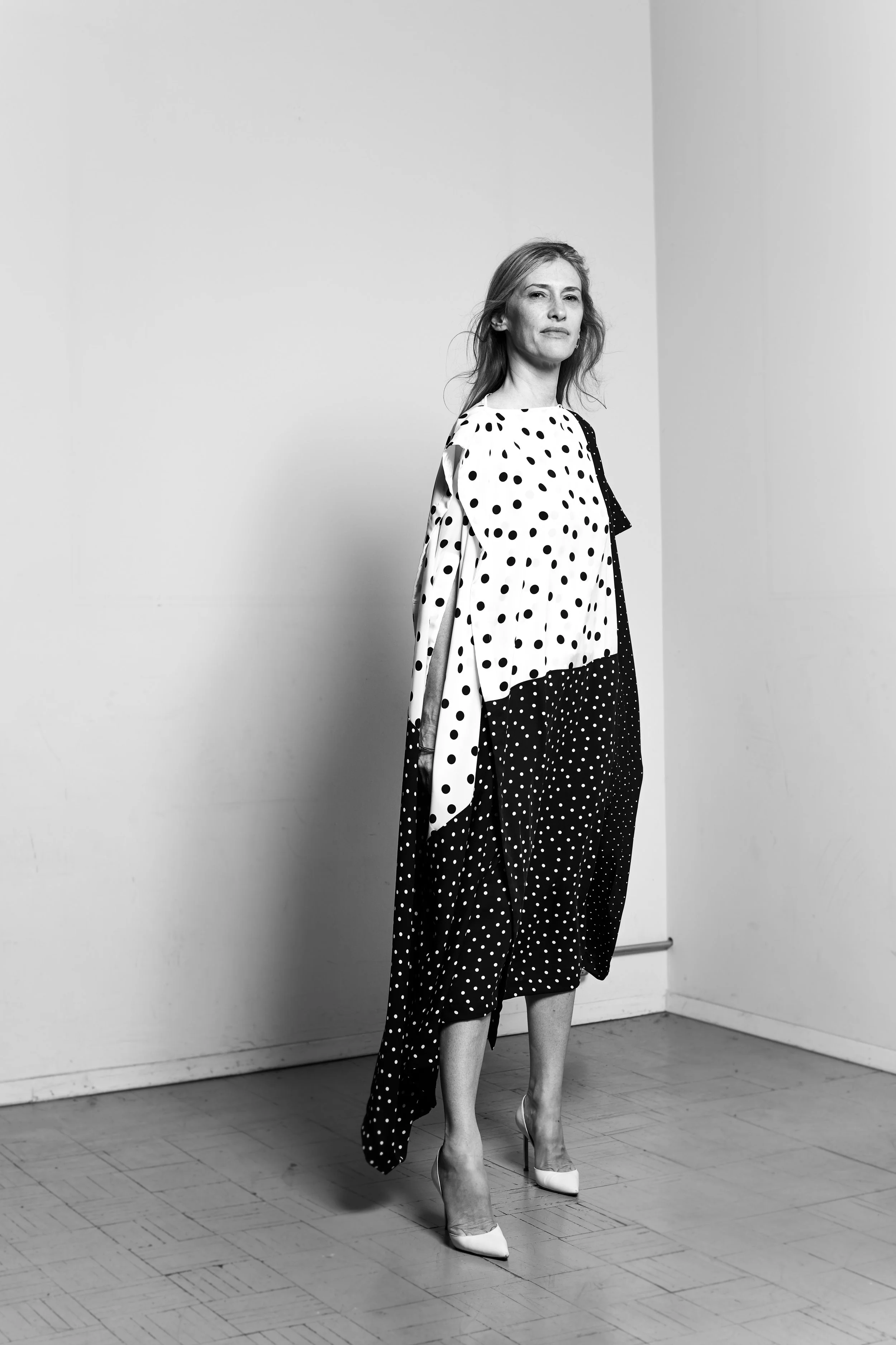 A woman with shoulder-length hair, wearing a flowing polka-dot dress with contrasting black and white sections, standing on a wooden floor in front of a plain wall with one corner visible.