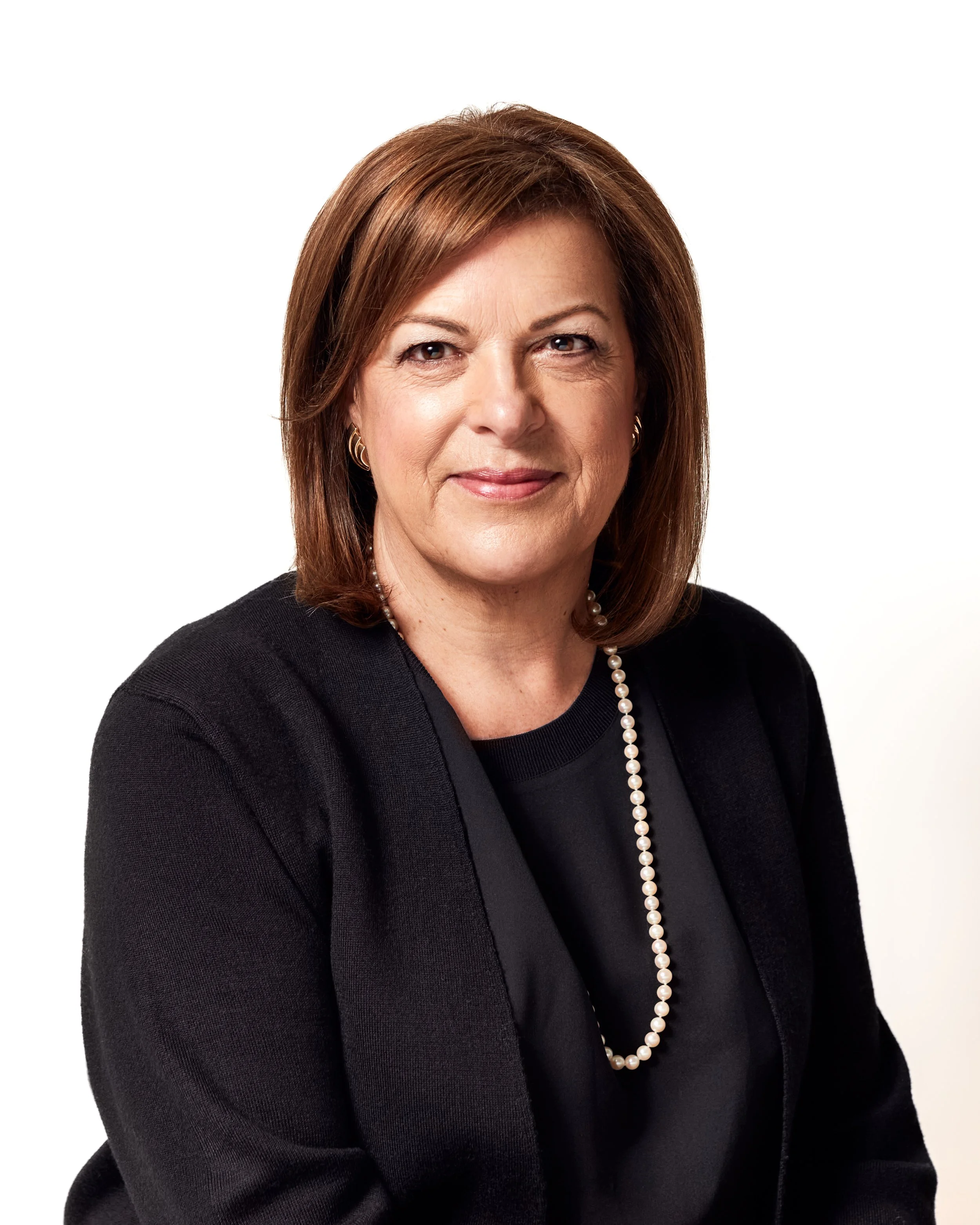 Headshot of a middle-aged woman with brown hair, wearing a black blazer and a pearl necklace, smiling slightly, against a plain white background.