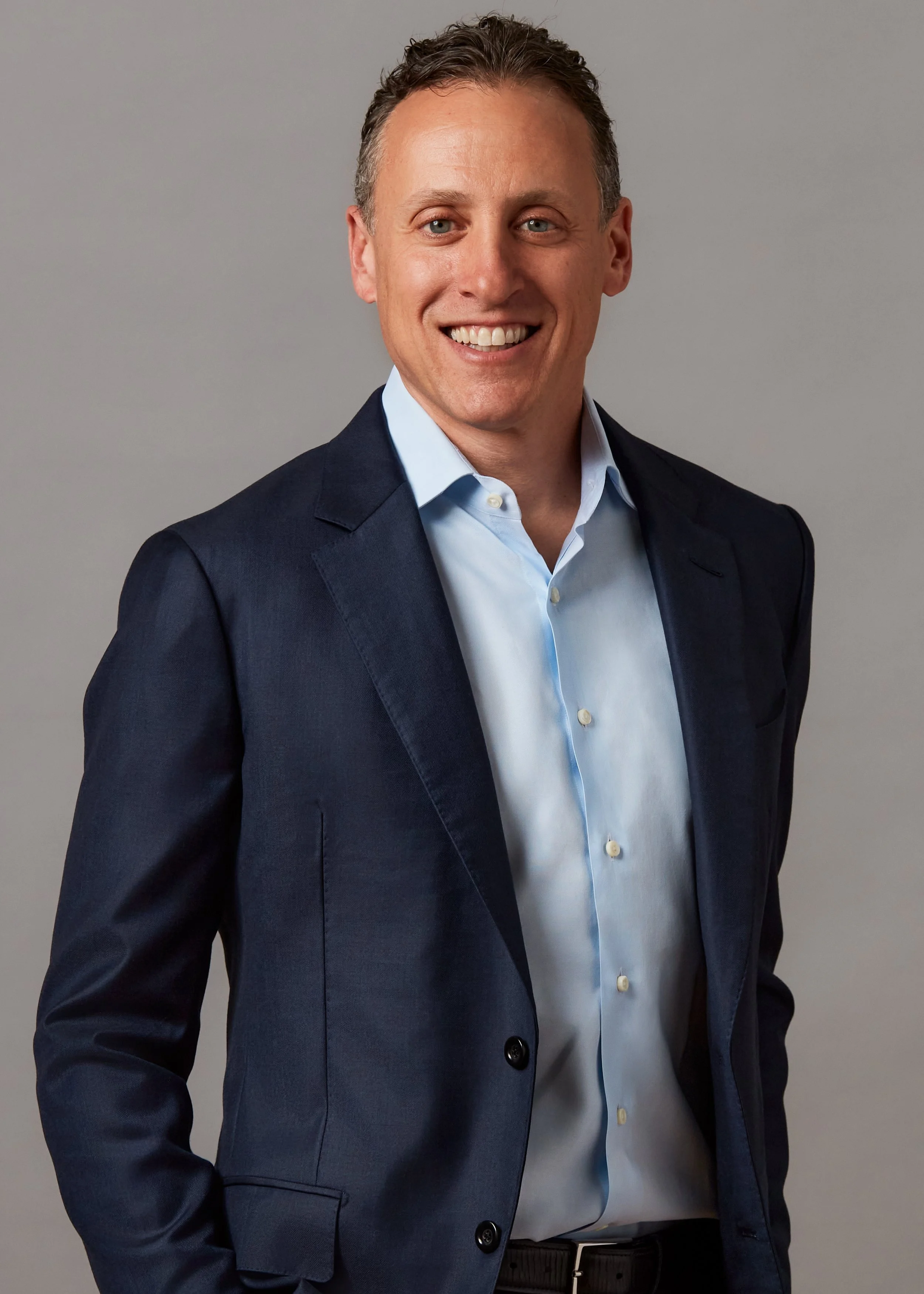 A man wearing a navy blue blazer and light blue shirt, smiling against a gray background.