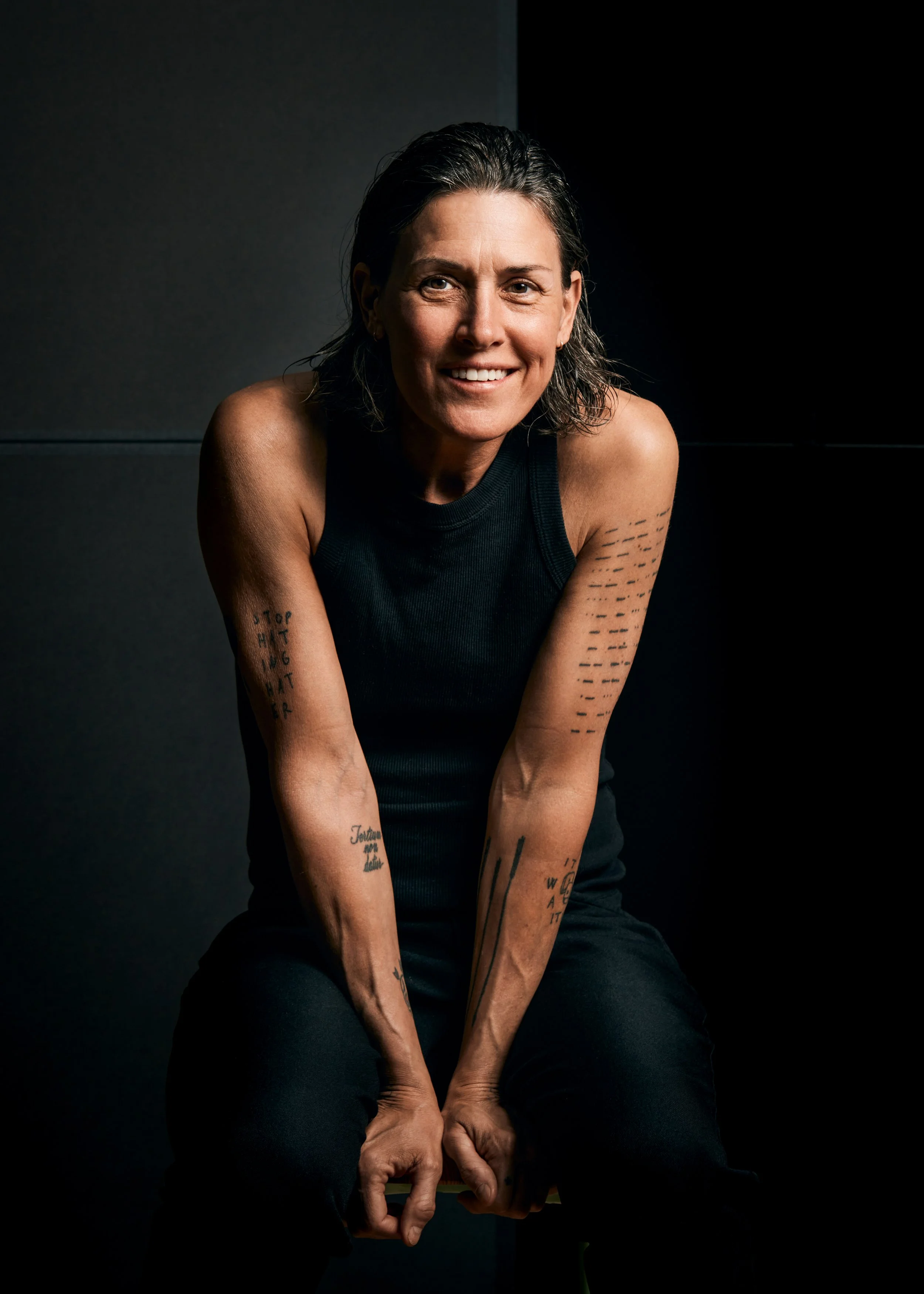 A smiling woman in a black sleeveless top, seated with her hands clasped between her legs, against a dark background. She has shoulder-length, dark, slightly wavy hair and tattoos on her arms.