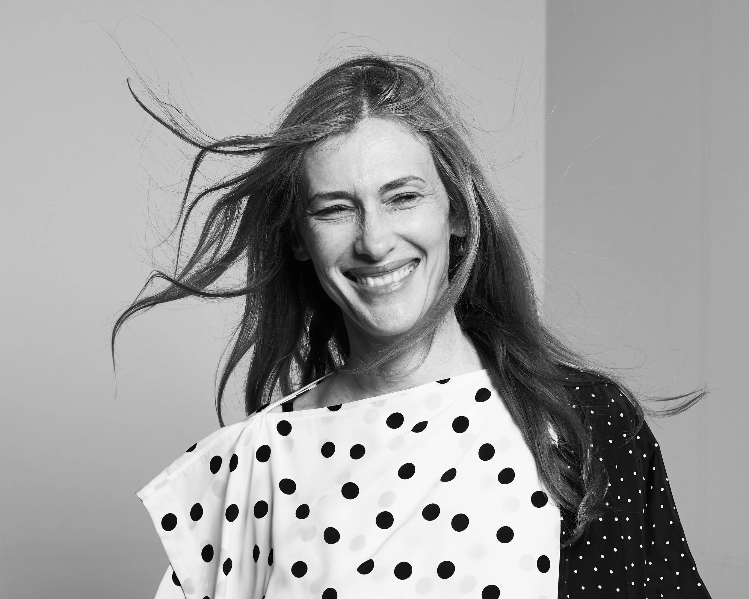 A woman with long hair smiling and winking, wearing a polka-dot dress and a contrasting polka-dot jacket.