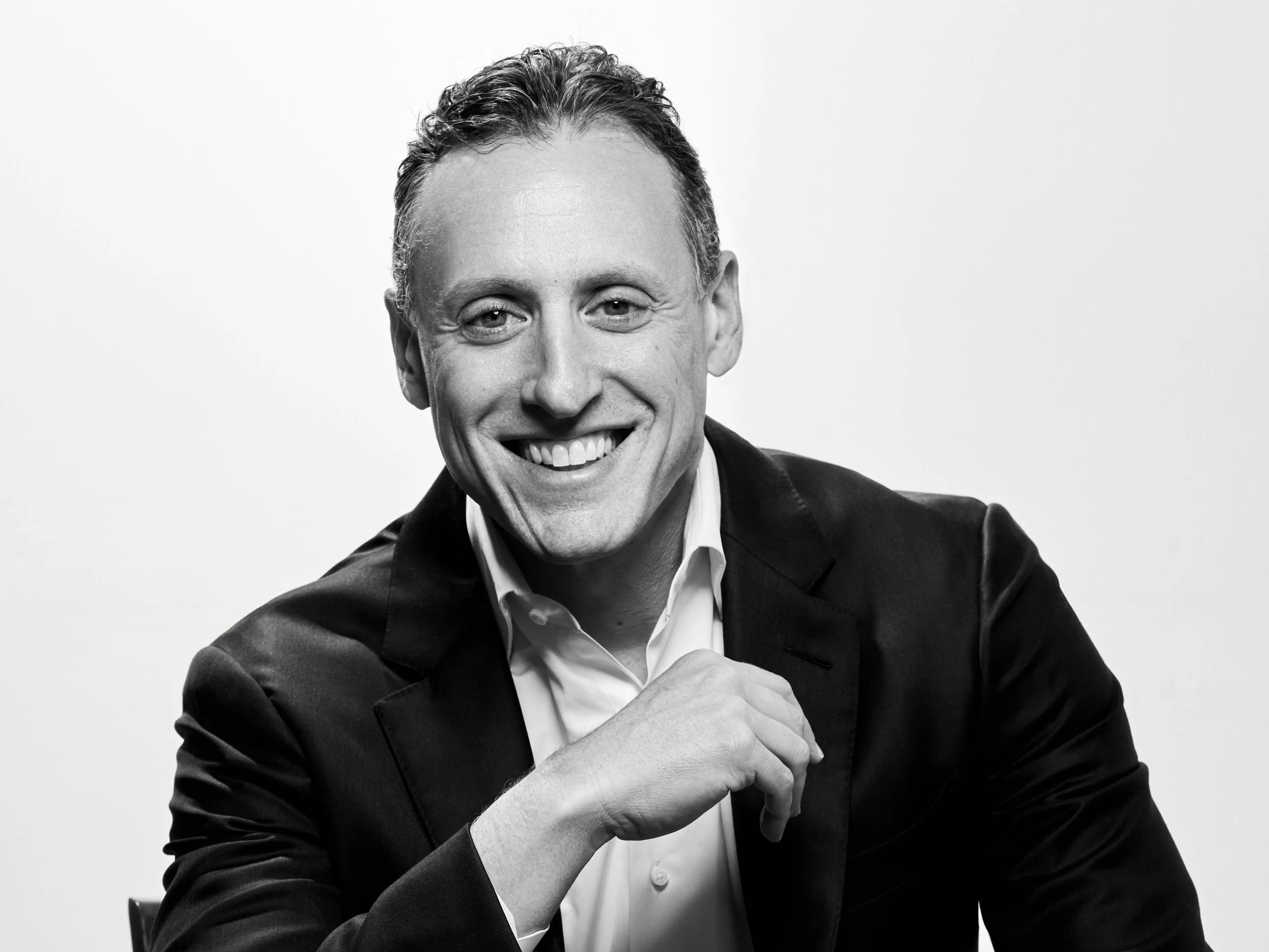 Black and white portrait of a smiling man in a suit and white shirt, posing with one hand near his chin against a plain background.