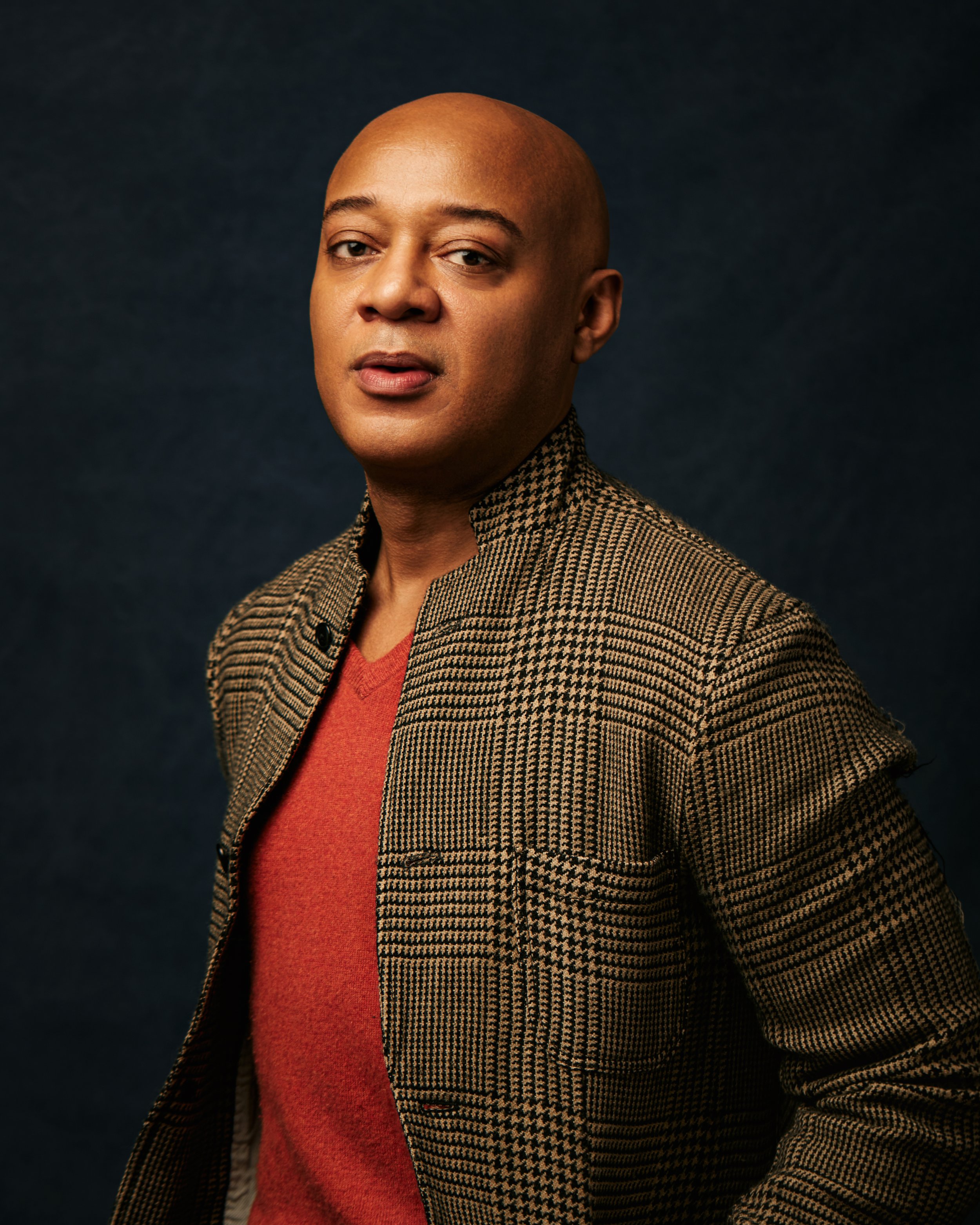 A man with a shaved head wearing a red shirt and a houndstooth blazer poses against a dark background.