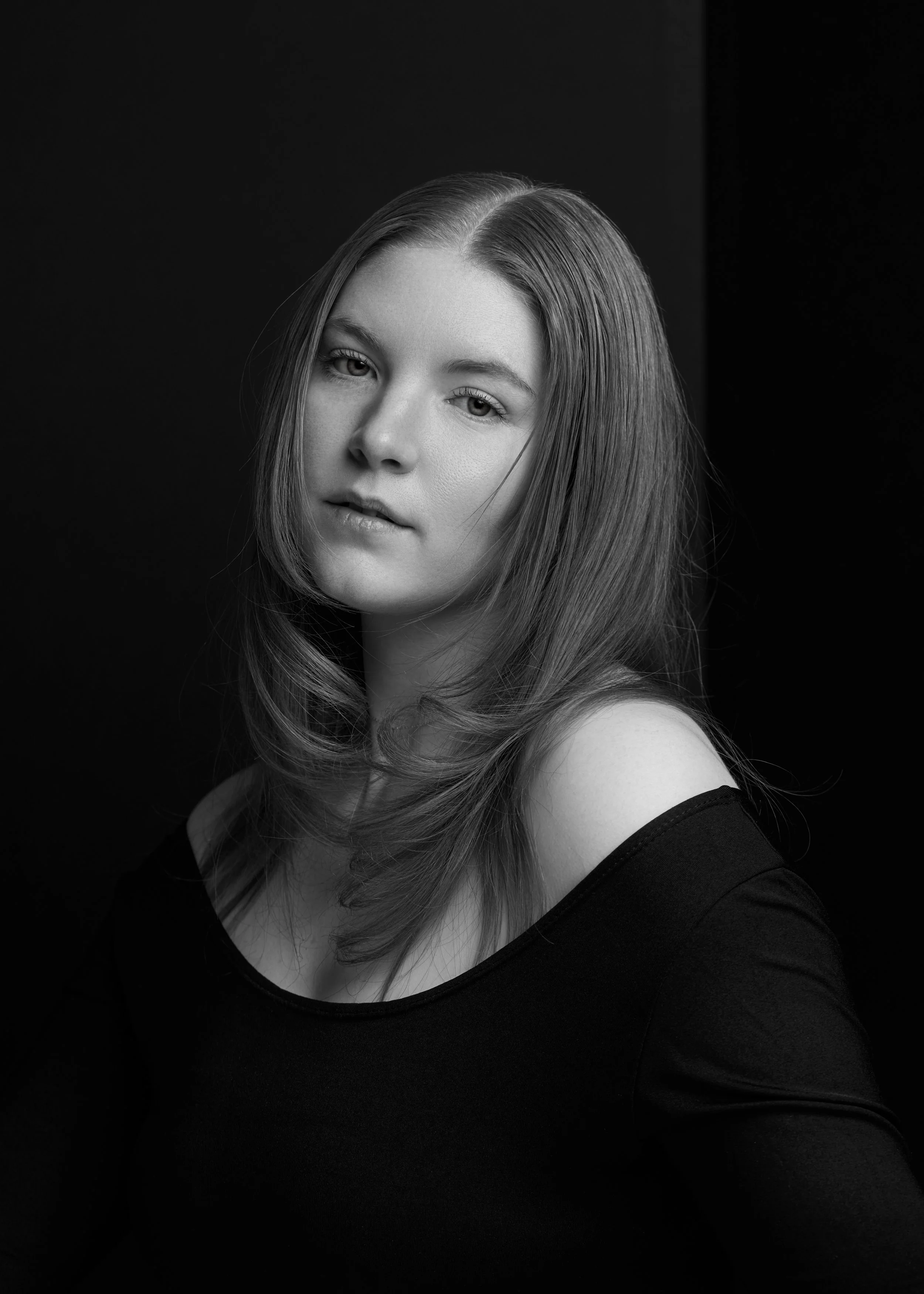 Black and white portrait of a young woman with long hair, wearing an off-the-shoulder top, with a neutral expression and slight head tilt, against a dark background.