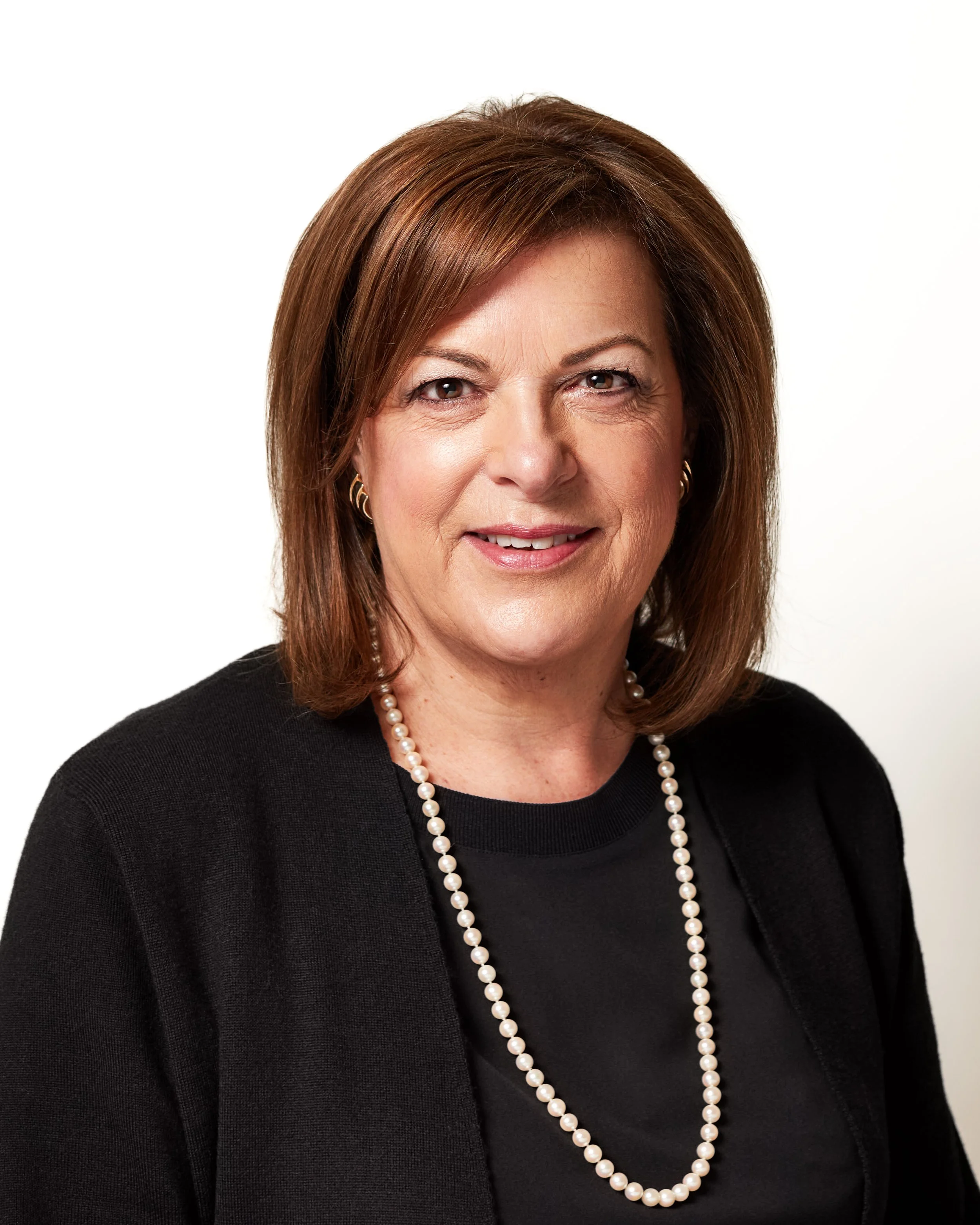 Portrait of a middle-aged woman with brown hair, wearing a black blazer, a black top, and a pearl necklace, smiling against a white background.