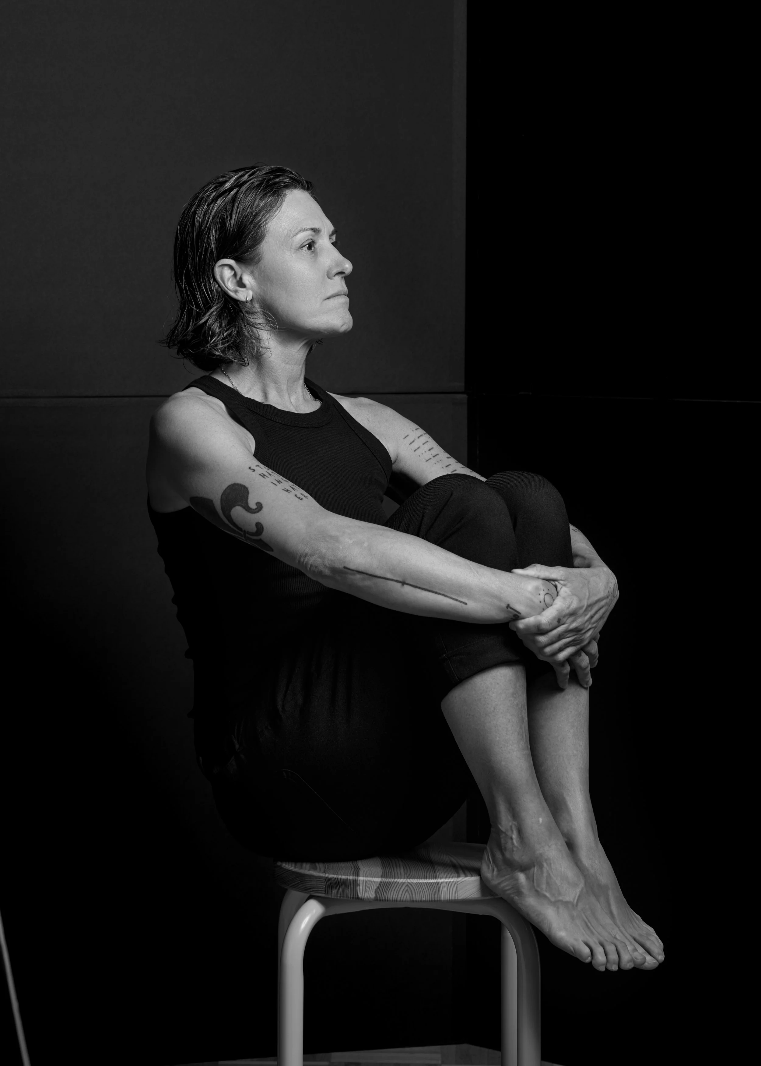 A black and white photograph of a woman with short hair sitting on a stool, holding her knees to her chest. She is wearing a sleeveless top and has tattoos on her arms. She appears to be deep in thought with a serious expression.