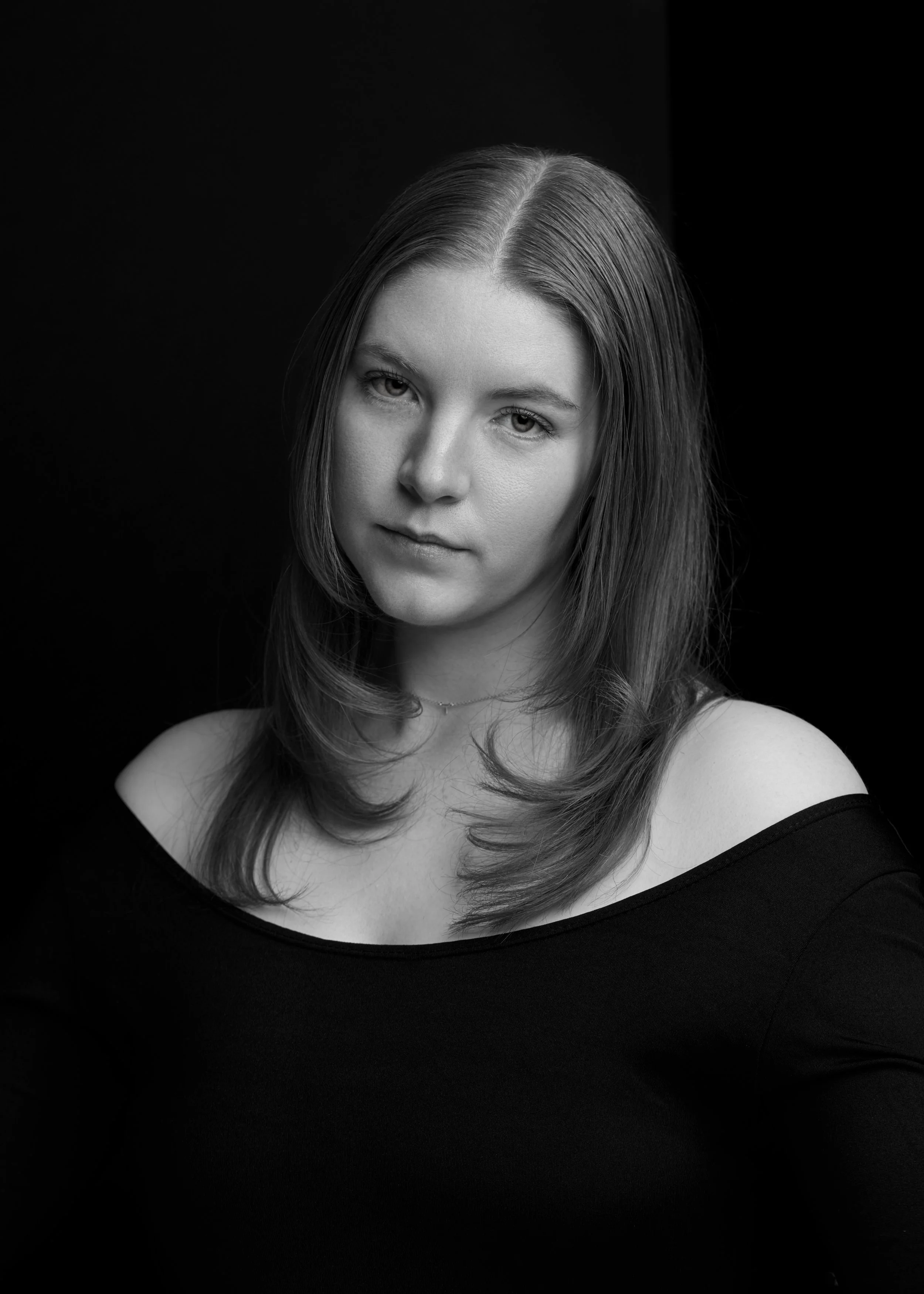 Black and white portrait of a young woman with shoulder-length hair, wearing an off-the-shoulder top, looking directly at the camera with a neutral expression.