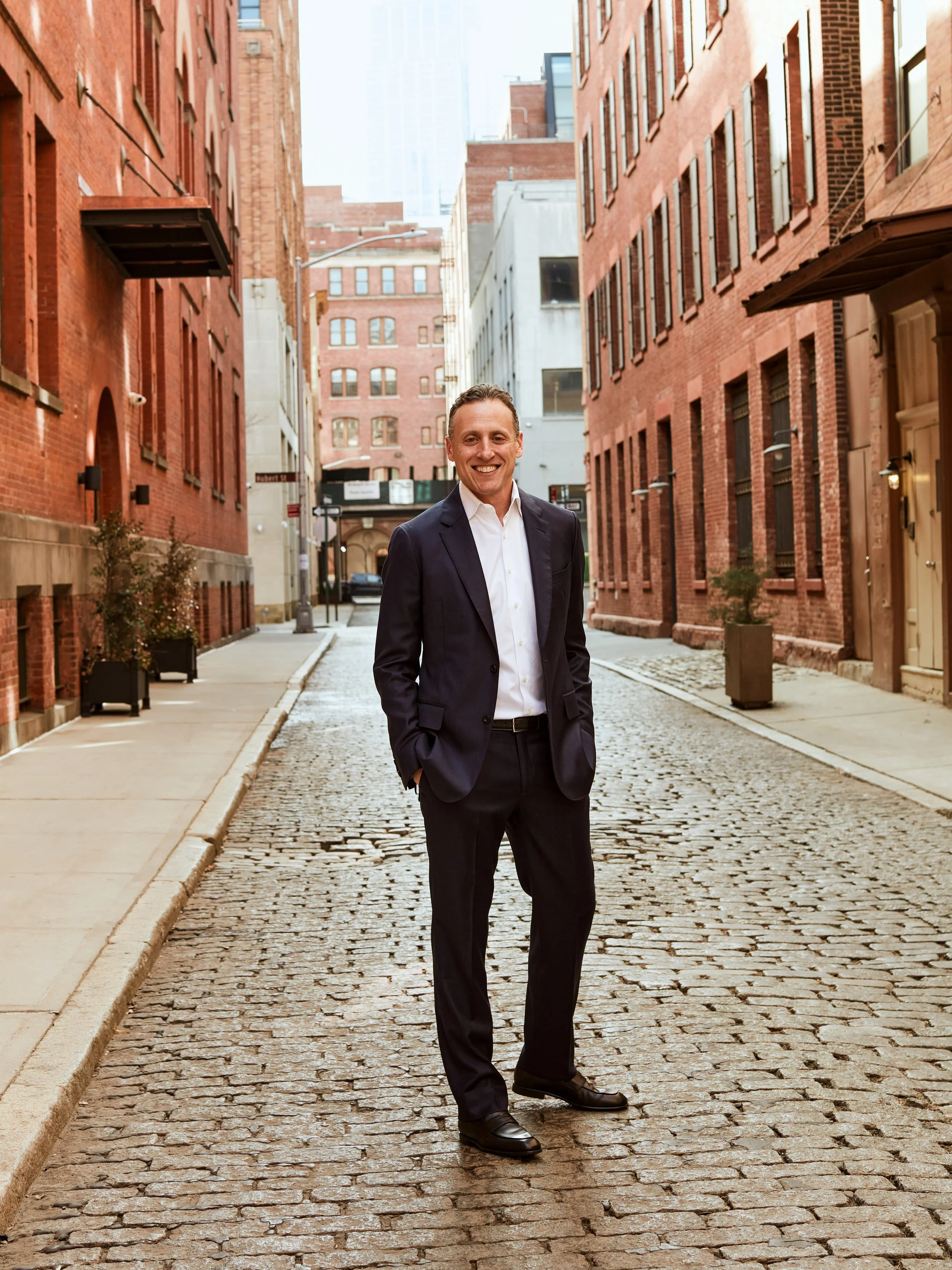 A man in a white shirt and dark suit standing on a cobblestone street in an urban area with brick buildings.