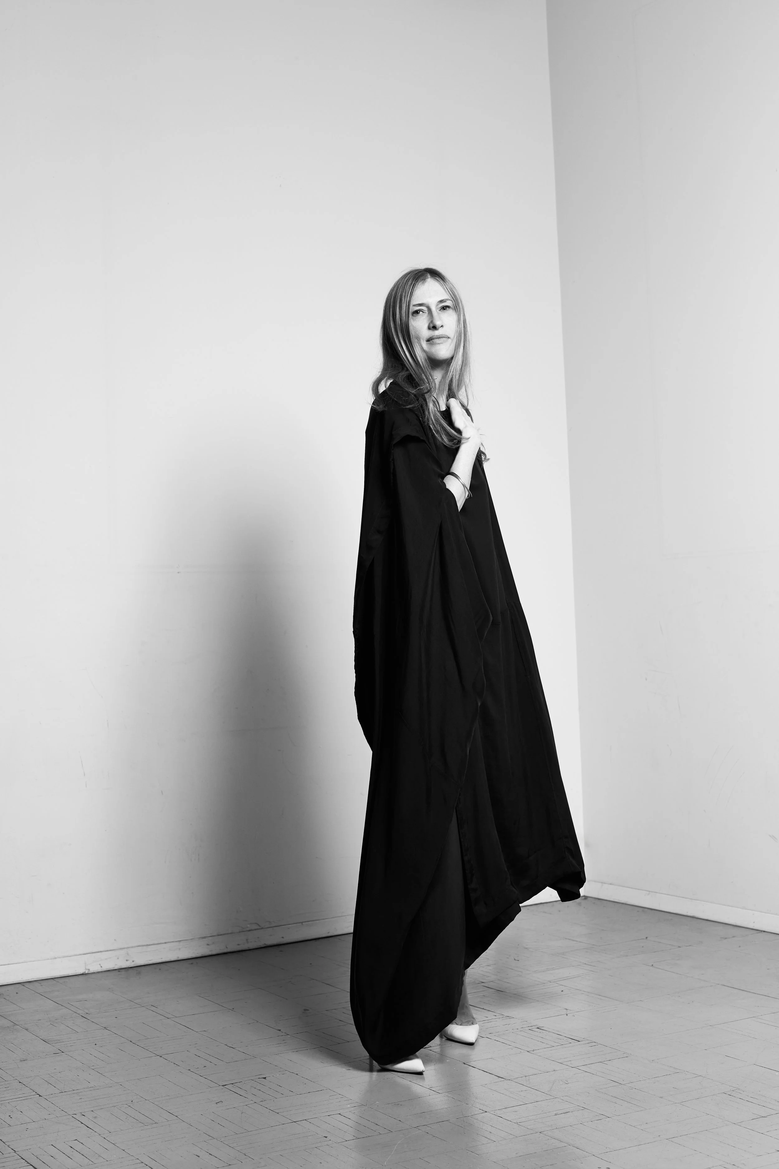 Black and white photo of a woman in a long dark dress and high heels, standing indoors against a plain wall.