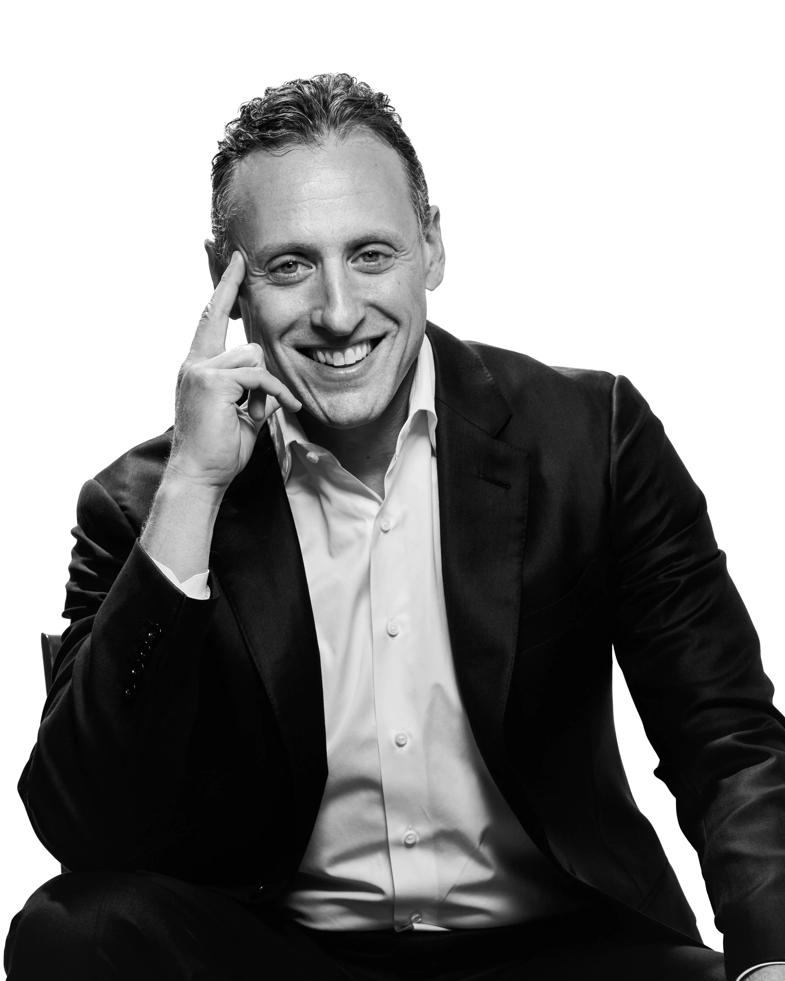 A black and white photo of a smiling man in a suit with a white shirt, sitting with his left arm resting on his knee and touching his temple with his right hand.