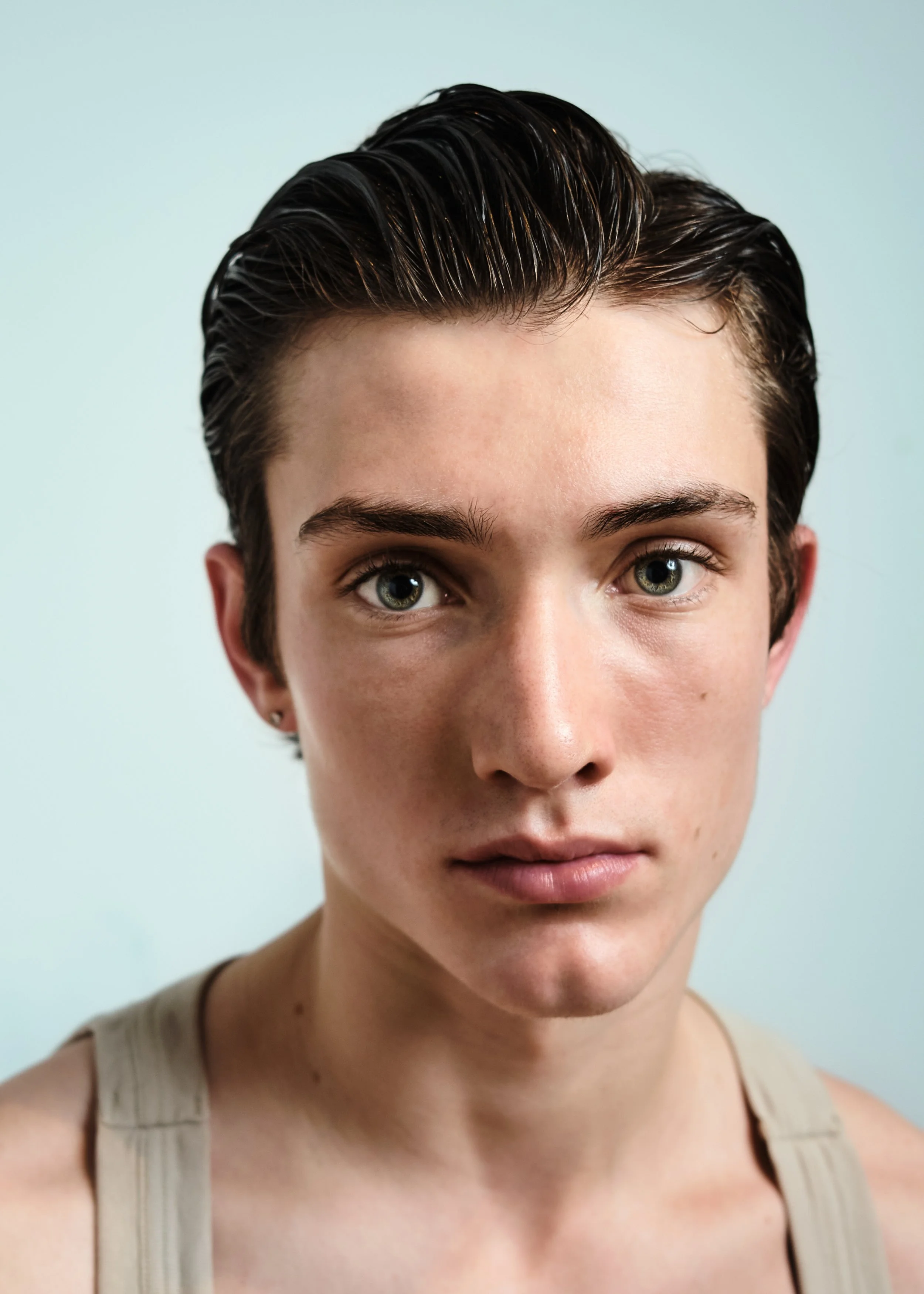 Close-up portrait of a young person with short, dark, slicked-back hair, fair skin, and striking eyes, wearing a beige tank top, against a light-colored background.