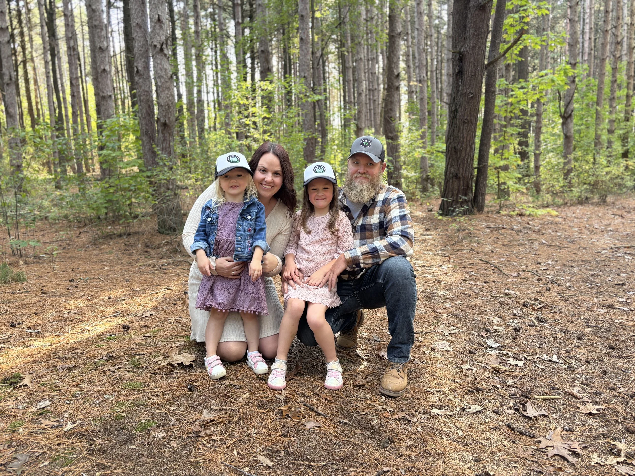Family of four, including mother, father, and two young girls, smiling and posing together in a forest during daytime, surrounded by tall trees and fallen leaves.